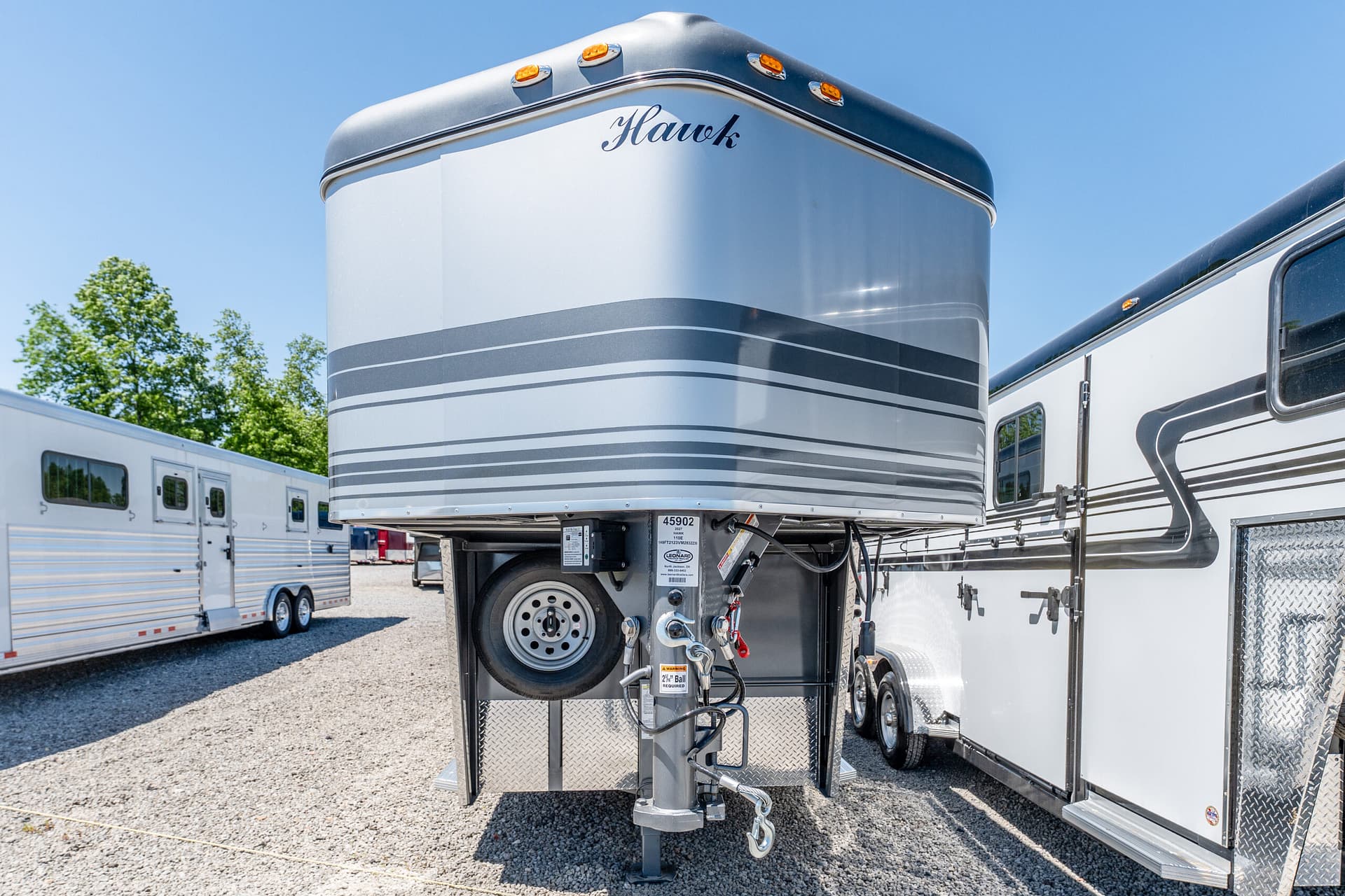 Gooseneck horse trailer in a gravel lot with front cap reading 'Hawk' and a spare tire beneath the hitch area.