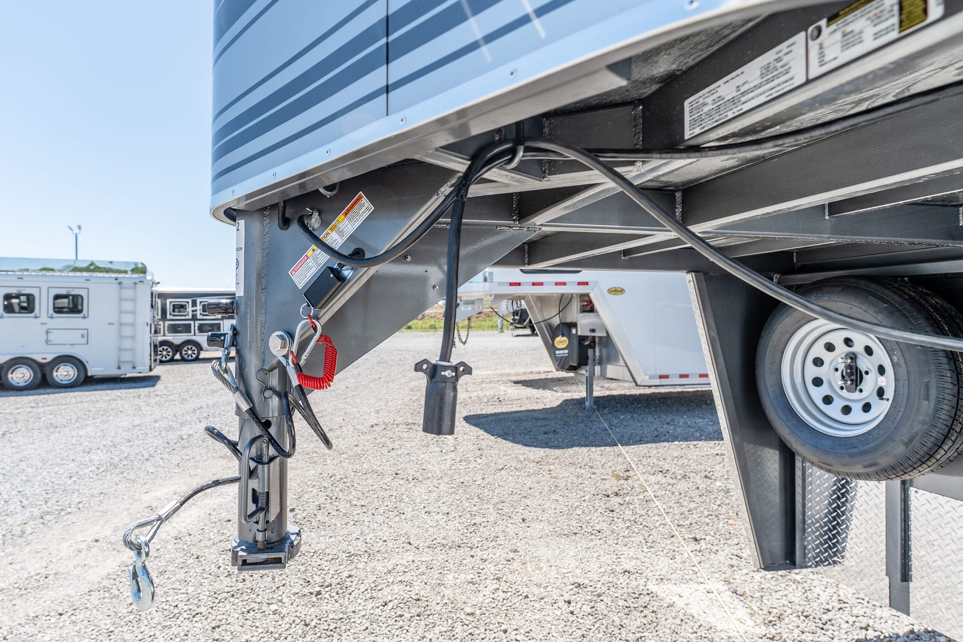 Close-up of the underside of a semi-trailer showing the hitch, cables, and a wheel on a gravel lot.