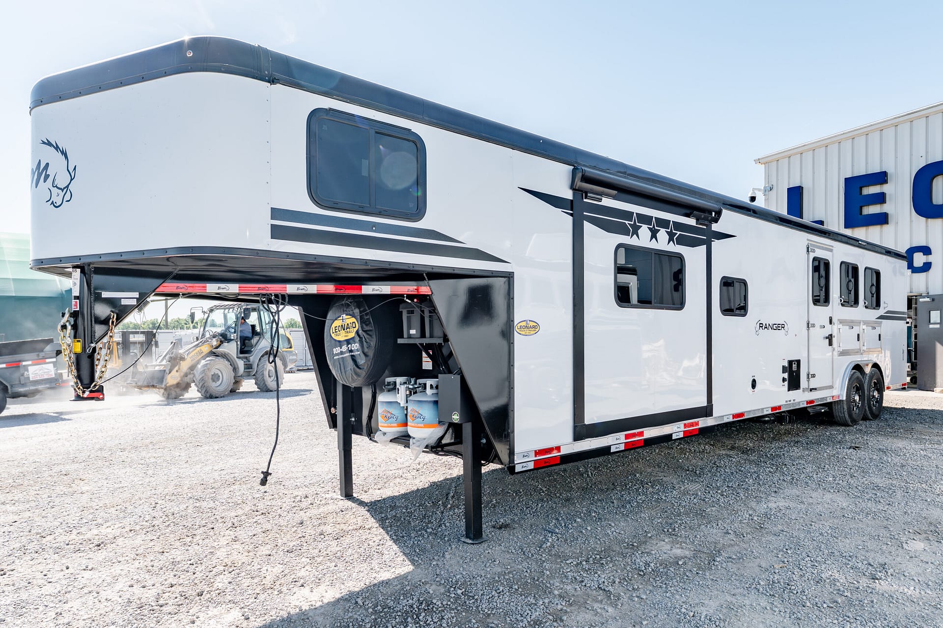 White fifth-wheel RV with blue-gray graphics parked on a gravel lot beside a service building; propane tanks beneath the hitch visible.