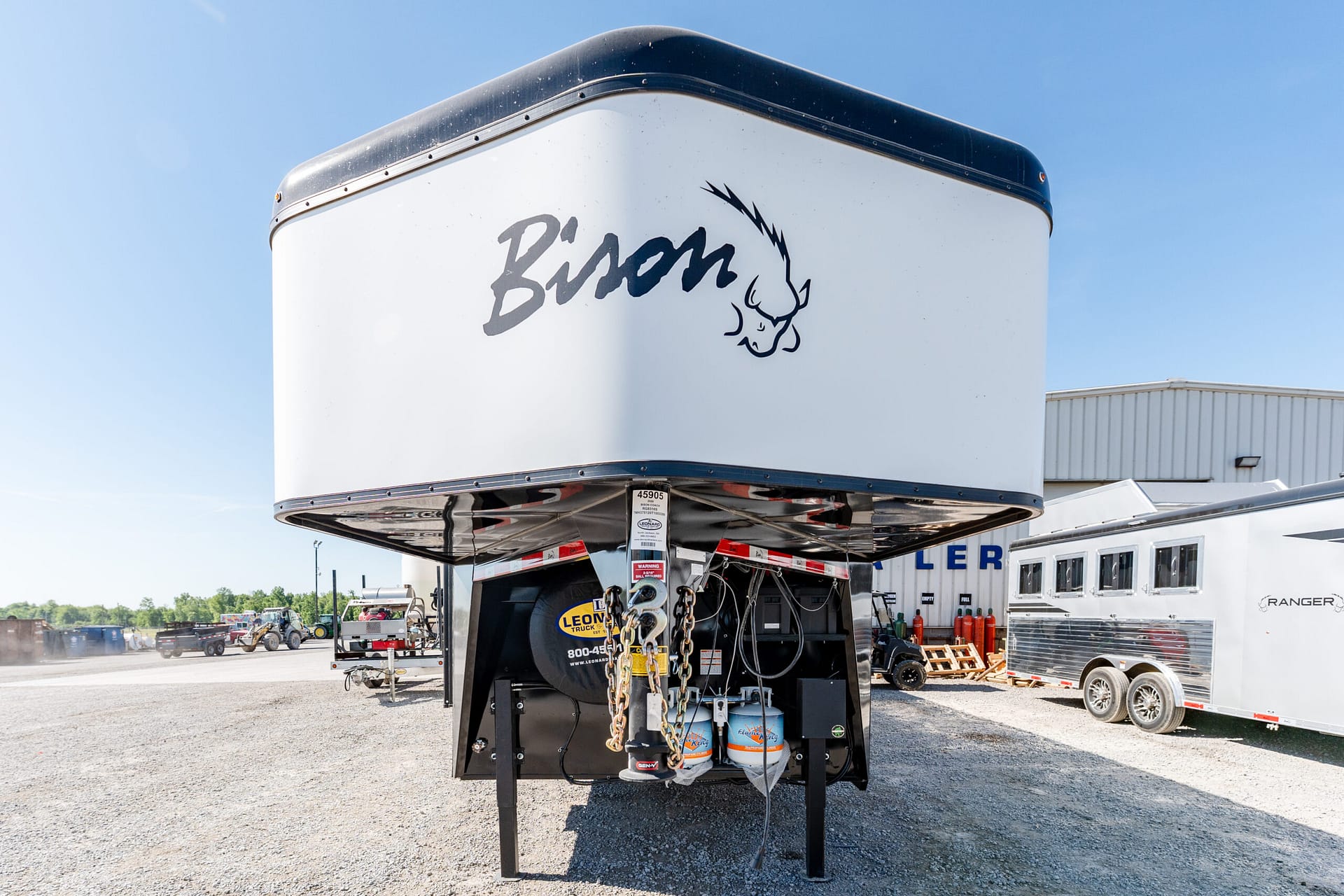 Rear of a white trailer with a dark blue top edge, displaying the word 'Boom' and a stylized bull head logo against a clear blue sky.