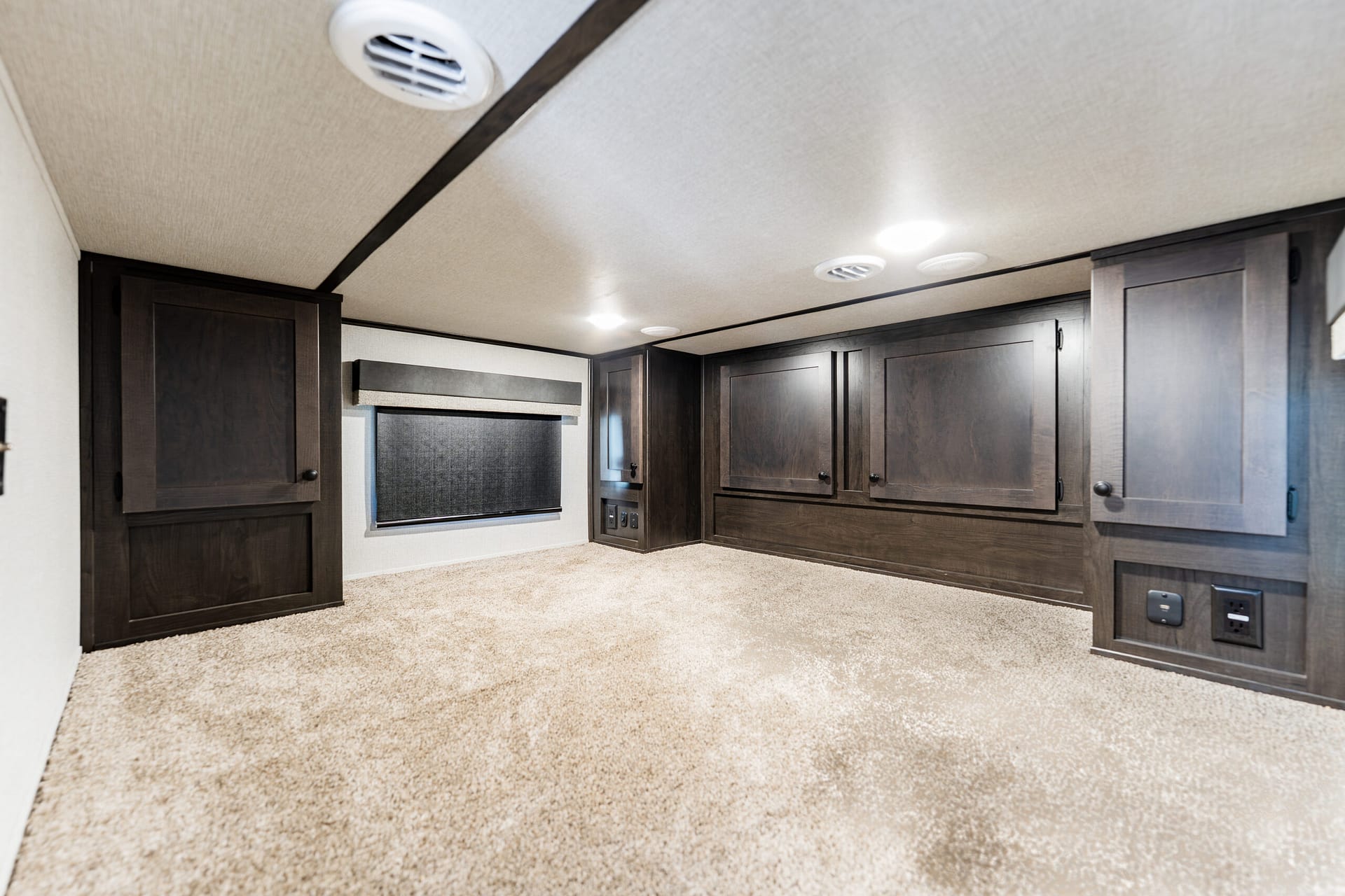 Interior of a room with dark wood cabinets along both walls, beige carpet, and a low ceiling with ceiling vents and lights, typical of an RV or trailer.