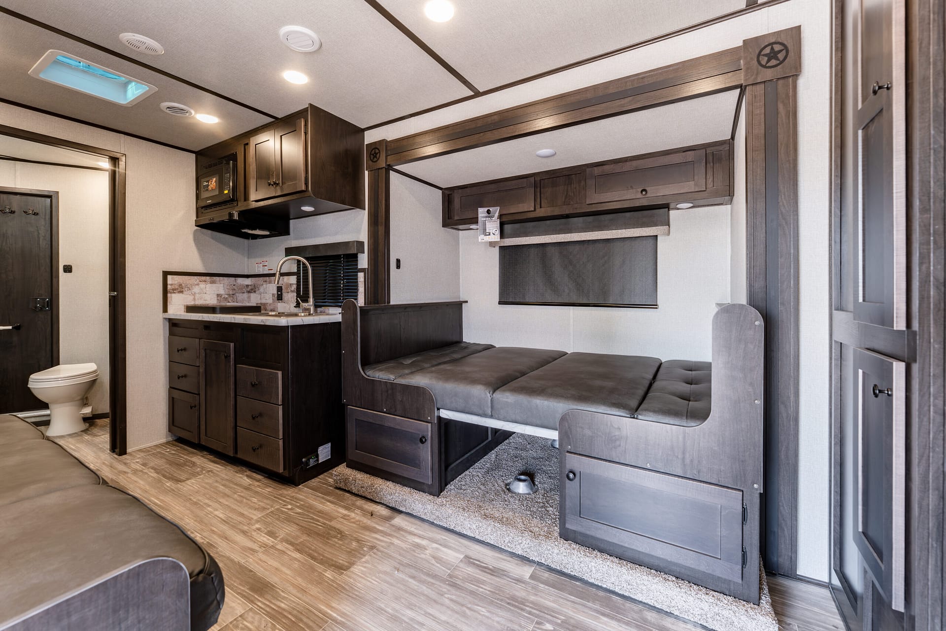 Interior of a compact RV kitchen and living area with dark wood cabinets, microwave, sink, and a cushioned sofa bed along the wall.