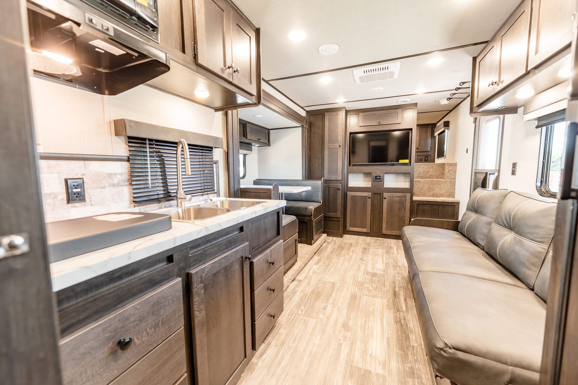 Interior of a modern RV kitchen and living area with dark wood cabinetry, granite counter, sink, and seating area with a sofa and TV.