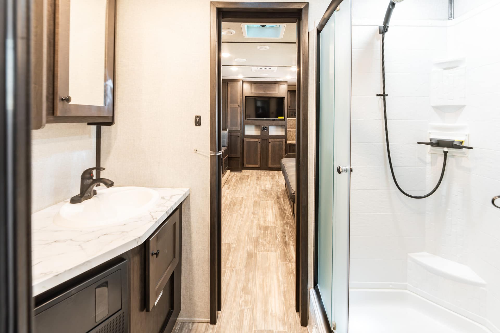 RV bathroom with marble countertop sink and dark wood cabinetry, hallway to living area outside the room in the distance with a TV visible in background, and a glass shower on the right.