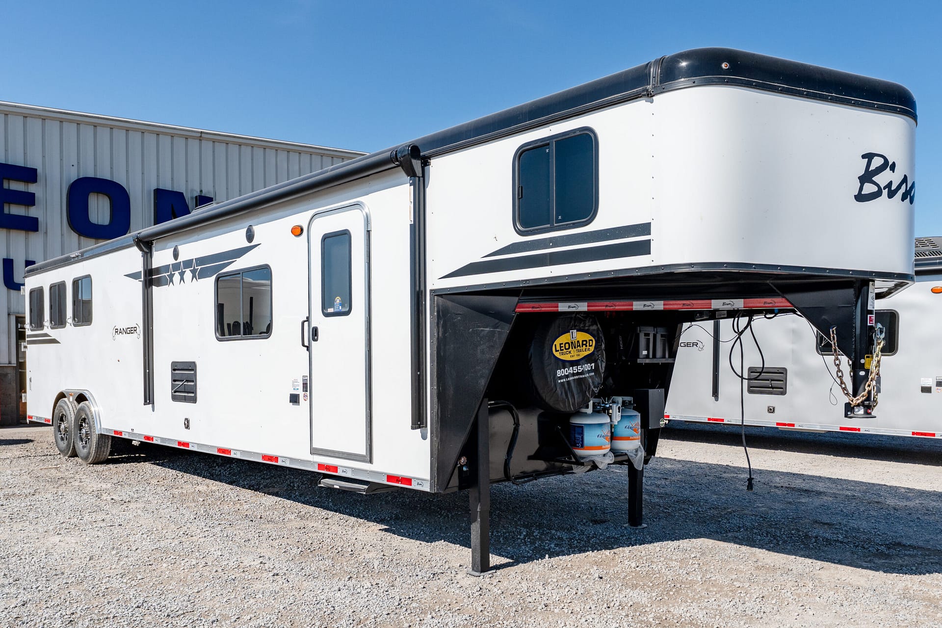 White fifth-wheel travel trailer with black trim parked on a gravel lot beside a building, stabilizing jacks deployed.