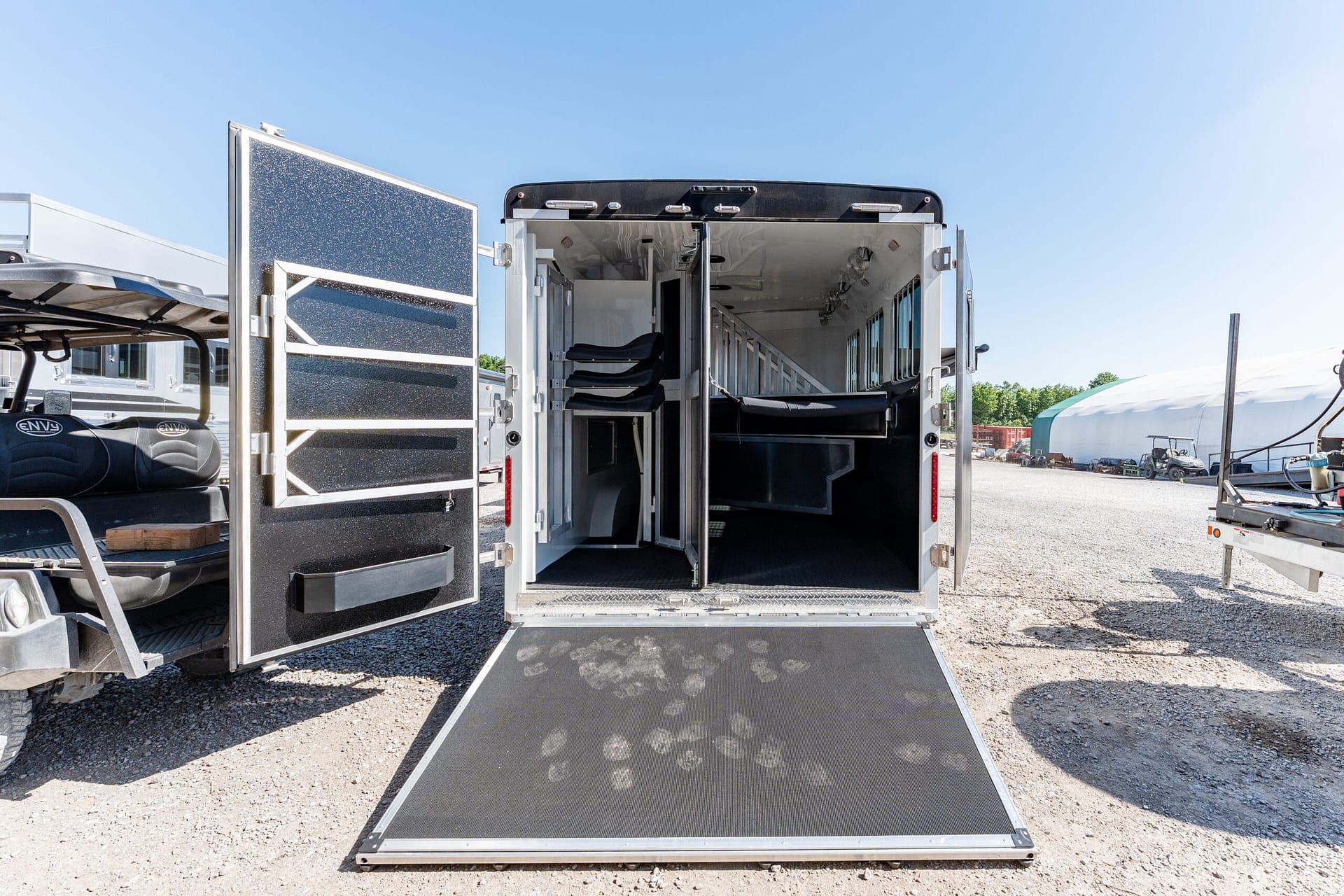 Open horse trailer with its rear doors swung wide and a ramp extended to the gravel, showing interior stalls and partitions inside under bright blue sky.
