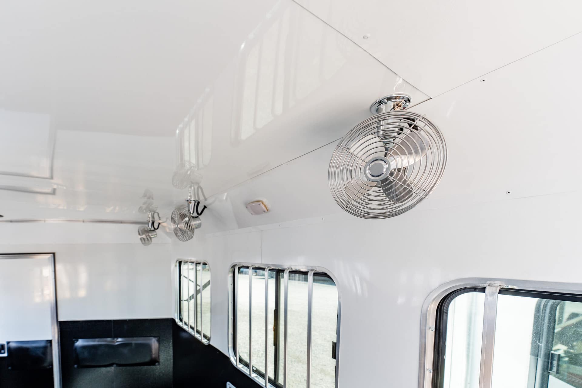 Interior of a white passenger vehicle with ceiling-mounted metal fans and a row of windows along one wall.