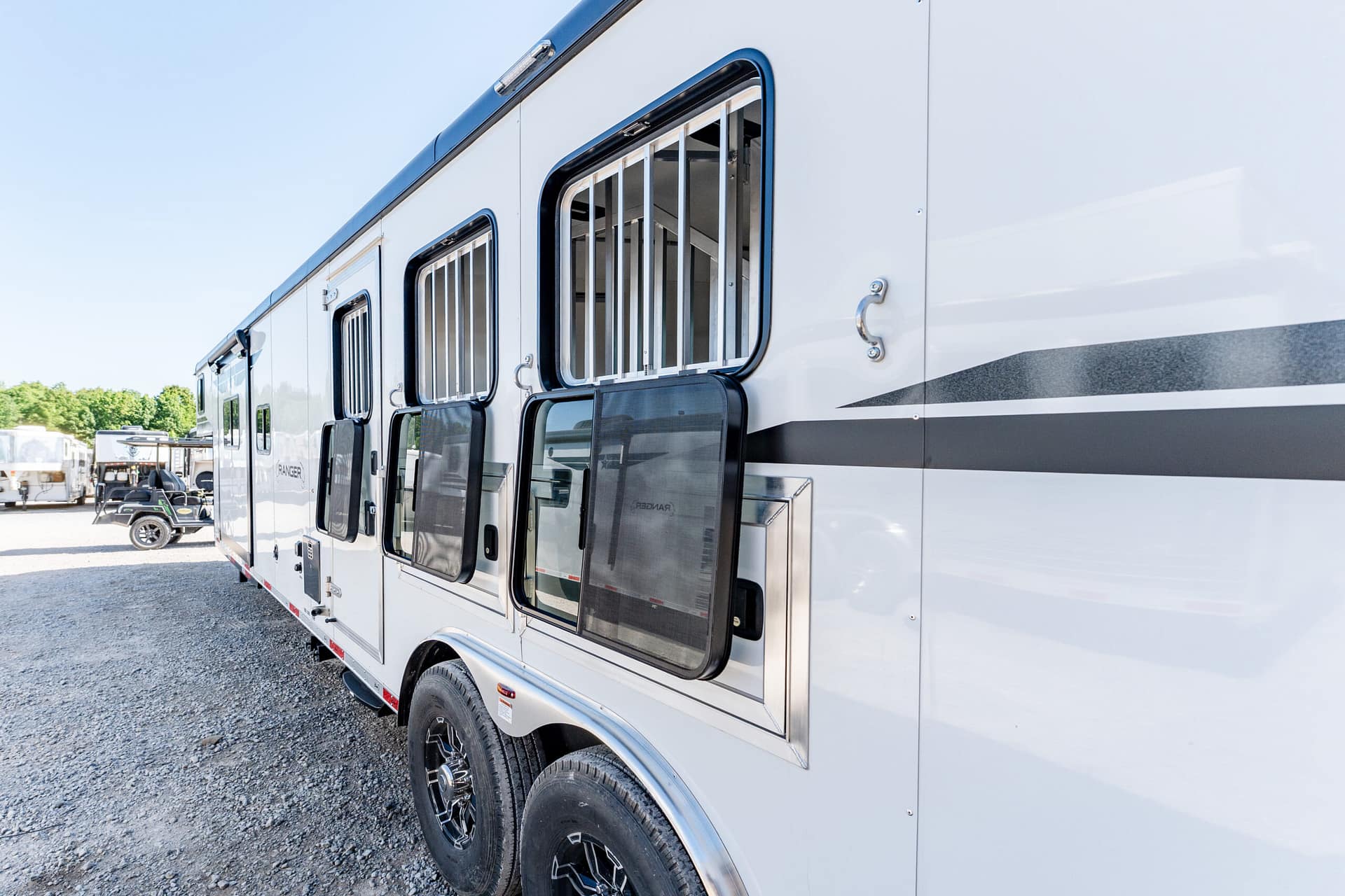 Side view of a silver RV with barred windows and a dark gray stripe, parked on a gravel lot.