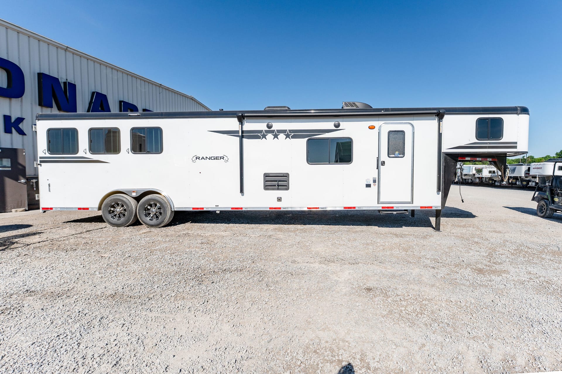White travel trailer parked on gravel in front of a warehouse under a bright blue sky, side view showing windows and dual axles.