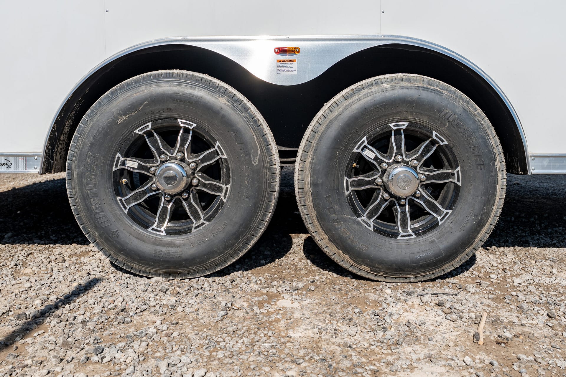 Two large trailer tires with chrome rims sit side by side on a gravel surface beneath a curved silver fender.