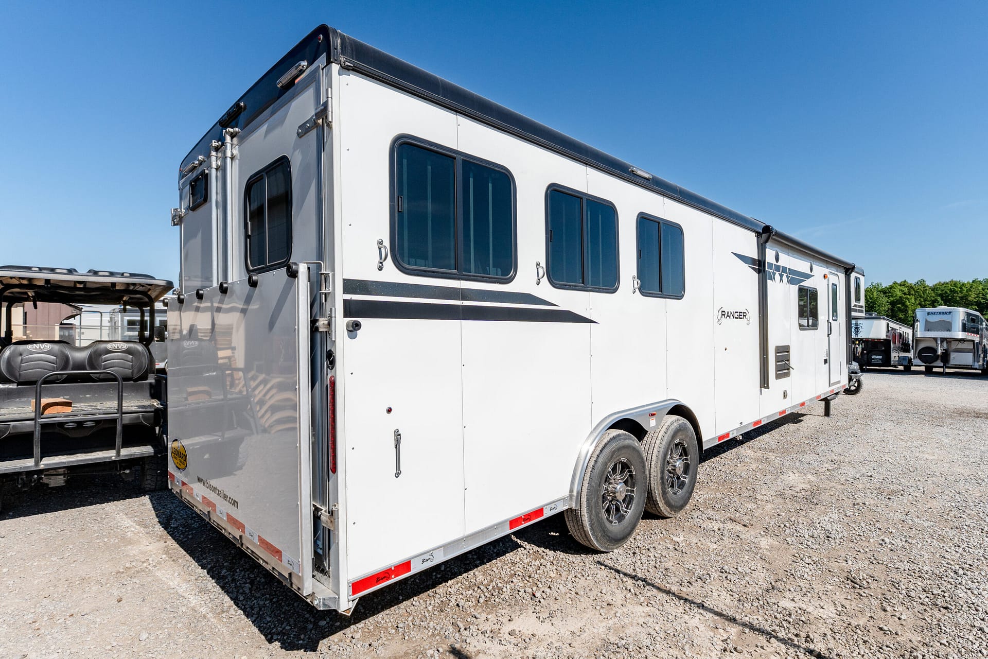 White horse trailer with multiple windows and black trim parked on a gravel lot under a clear blue sky, with other trailers visible in the background