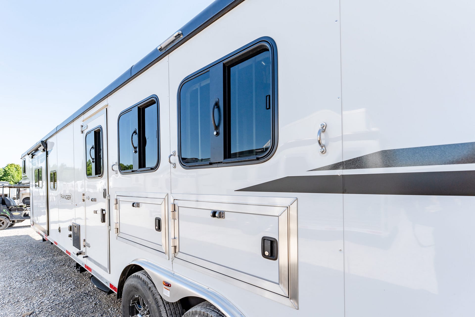 Side view of a white Ranger utility trailer with multiple small windows, external storage compartments, and a gray decorative stripe against a clear sky.