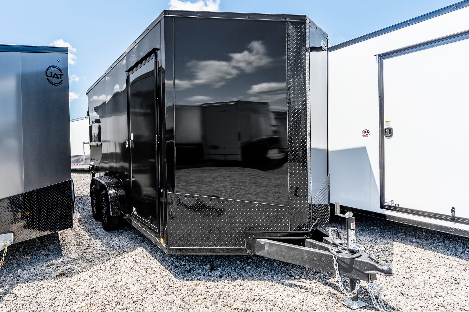 Black enclosed cargo trailer with diamond-plate front, hitched to a trailer in a gravel lot under a blue sky, flanked by other trailers.