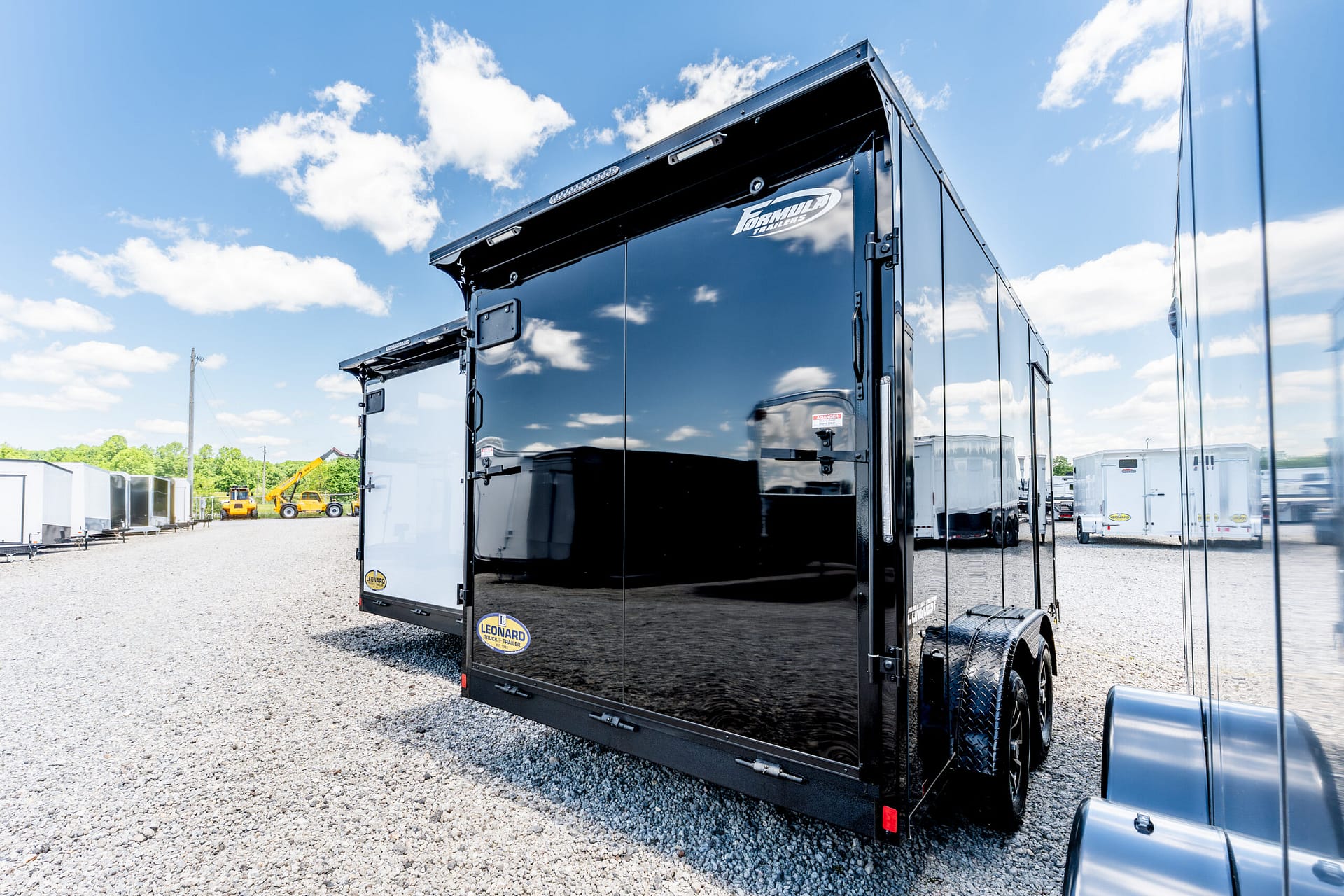 Black glossy trailer parked on a gravel lot with a clear blue sky and white clouds, reflecting surroundings.