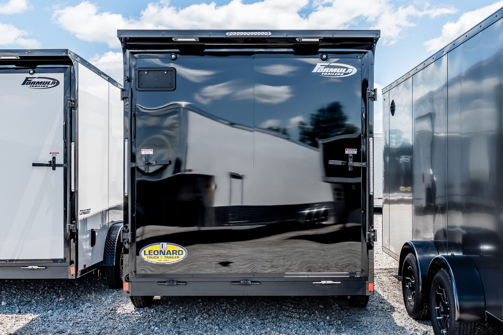 Rear view of a glossy black trailer with a Leonard sticker at the bottom center, flanked by white and gray trailers under a blue sky with clouds.