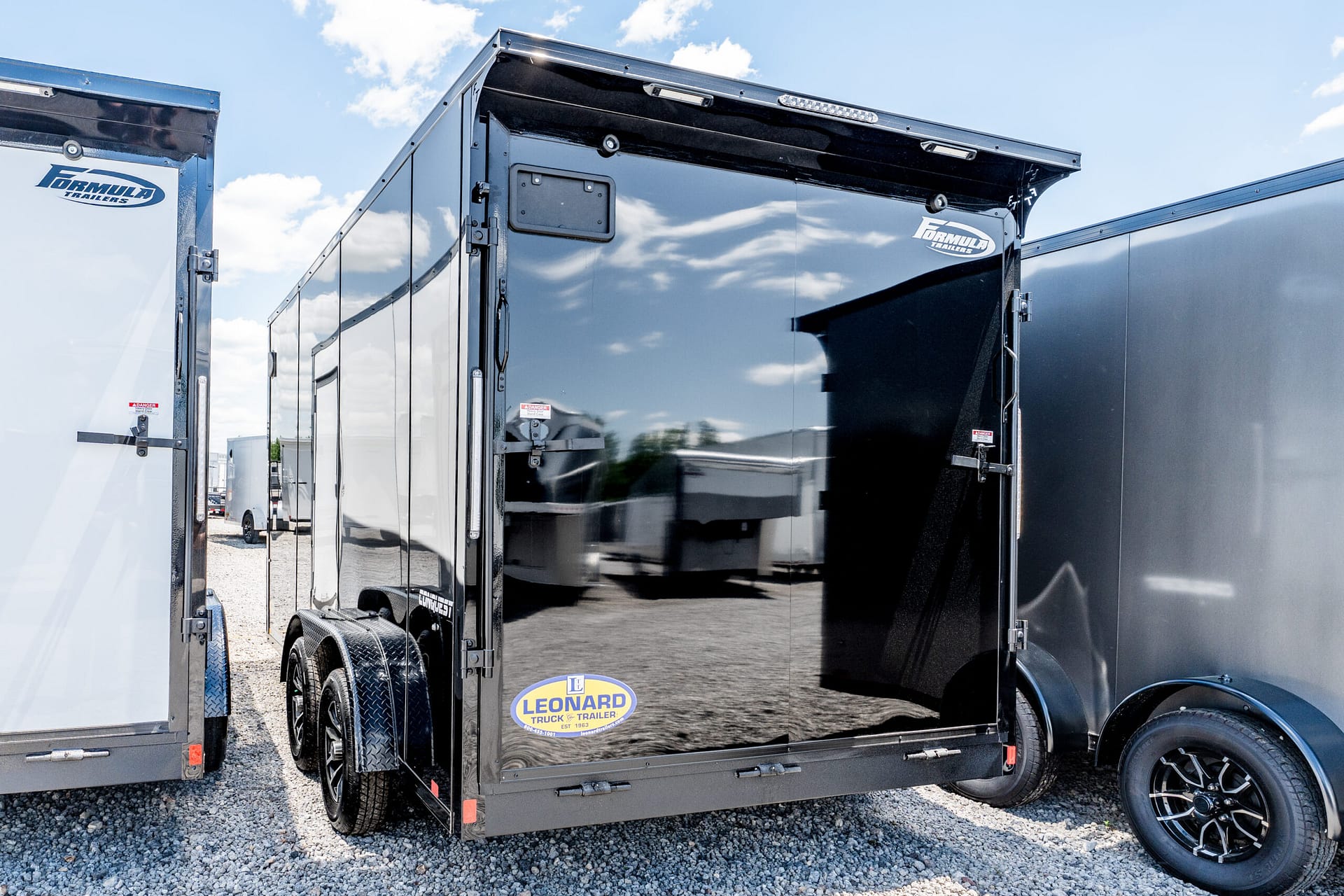 Shiny black enclosed trailer with a side door, reflections on the surface, and a Leonard Truck & Trailer logo near the bottom edge in a gravel lot