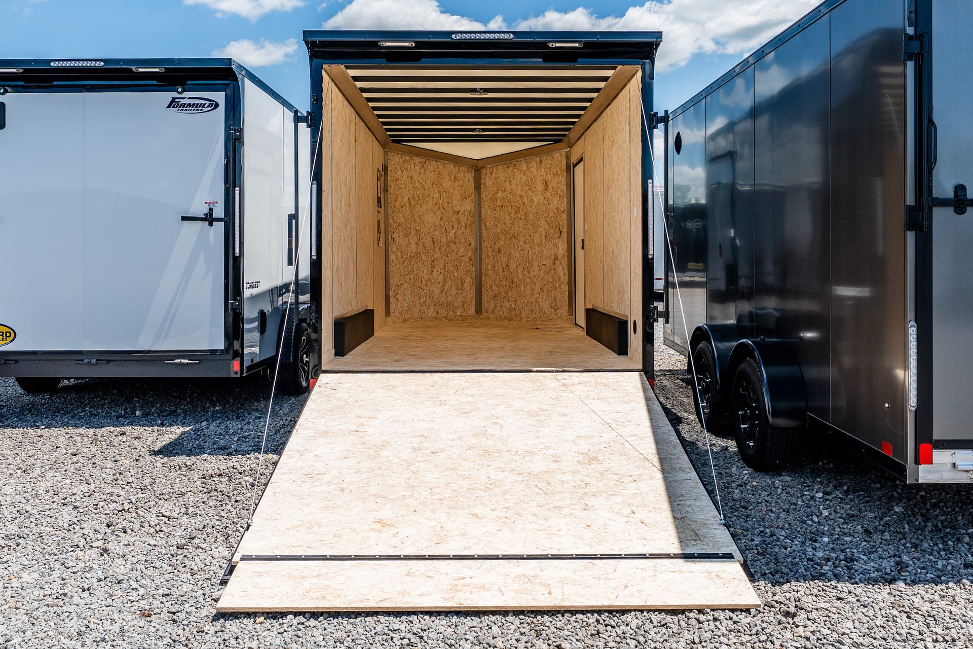 Open cargo trailer with wooden interior and ramp down, parked between two other trailers on a gravel lot.