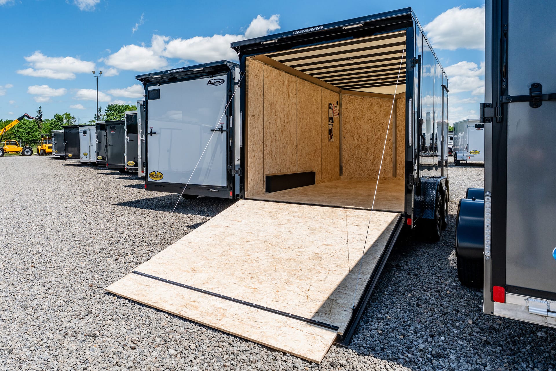 Open cargo trailer with ramp down, plywood interior, and other trailers on a gravel lot under a blue sky with white clouds.