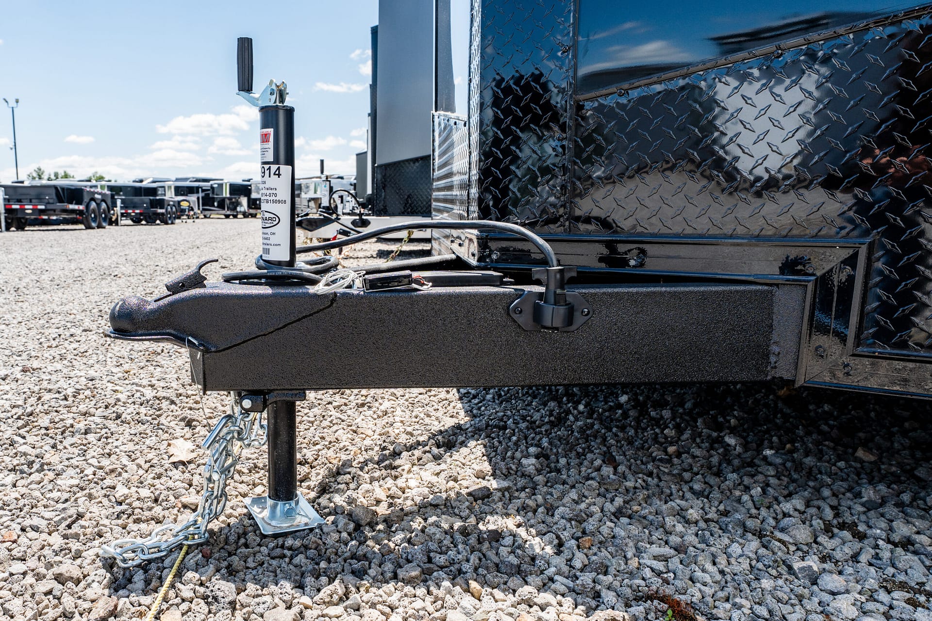 Close-up of a trailer tongue with a vertical jack and safety chains on a gravel lot, next to a shiny diamond-plate trailer side edge.