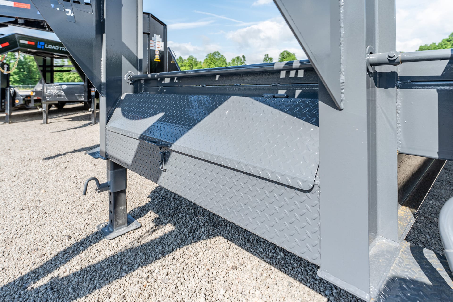 Close-up of gray steel industrial equipment with diamond-plate steps and supporting beams on a gravel lot (work trailer or lift component)