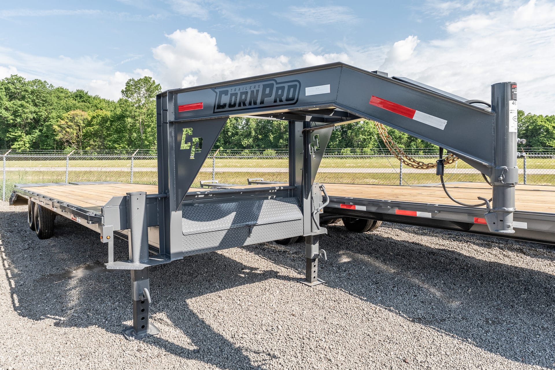 Gooseneck flatbed trailer painted gray on a gravel lot, with safety chains and a chain-link fence in the background and trees beyond.