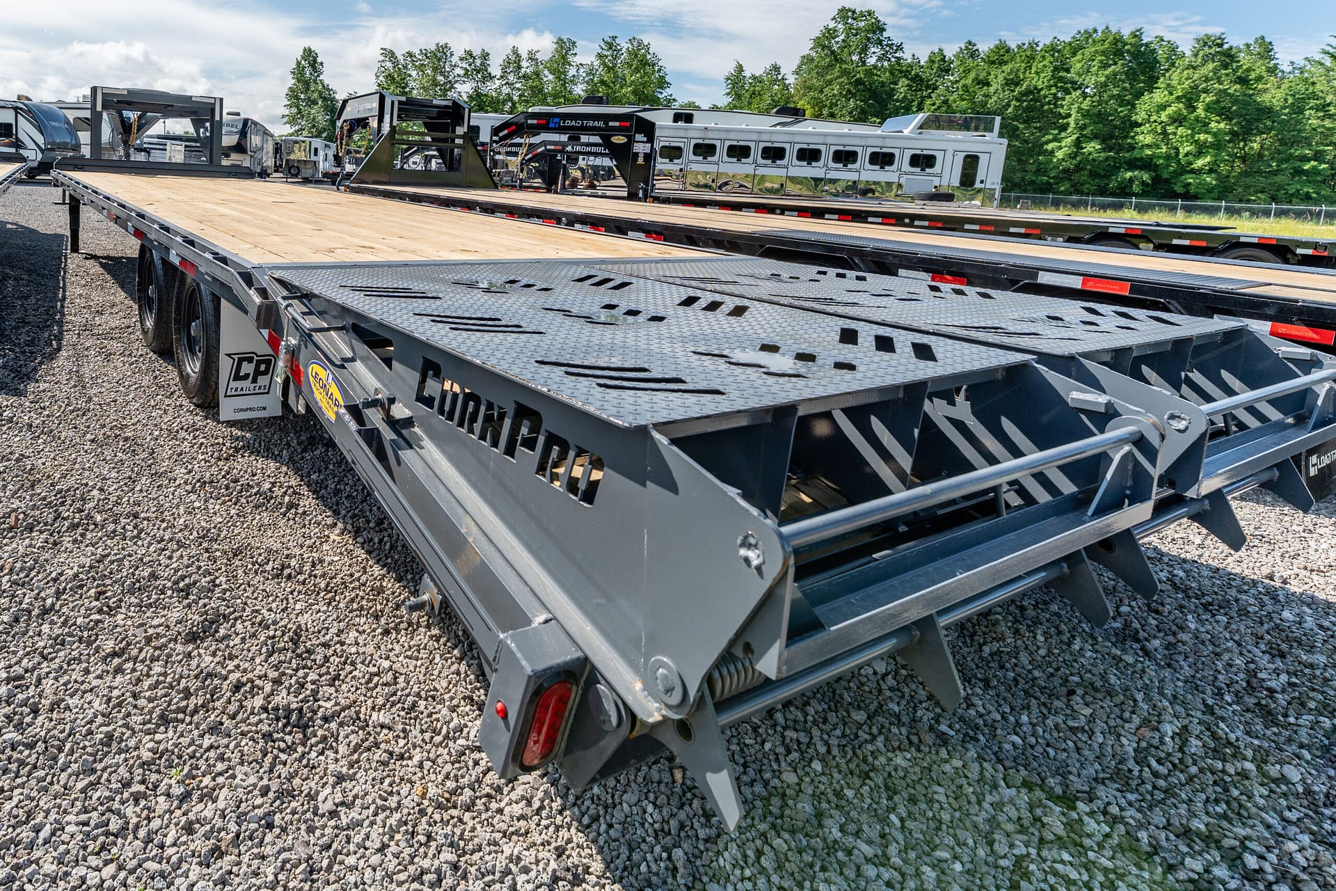 Flatbed trailer on a gravel lot with other trailers and a line of trees in the background.