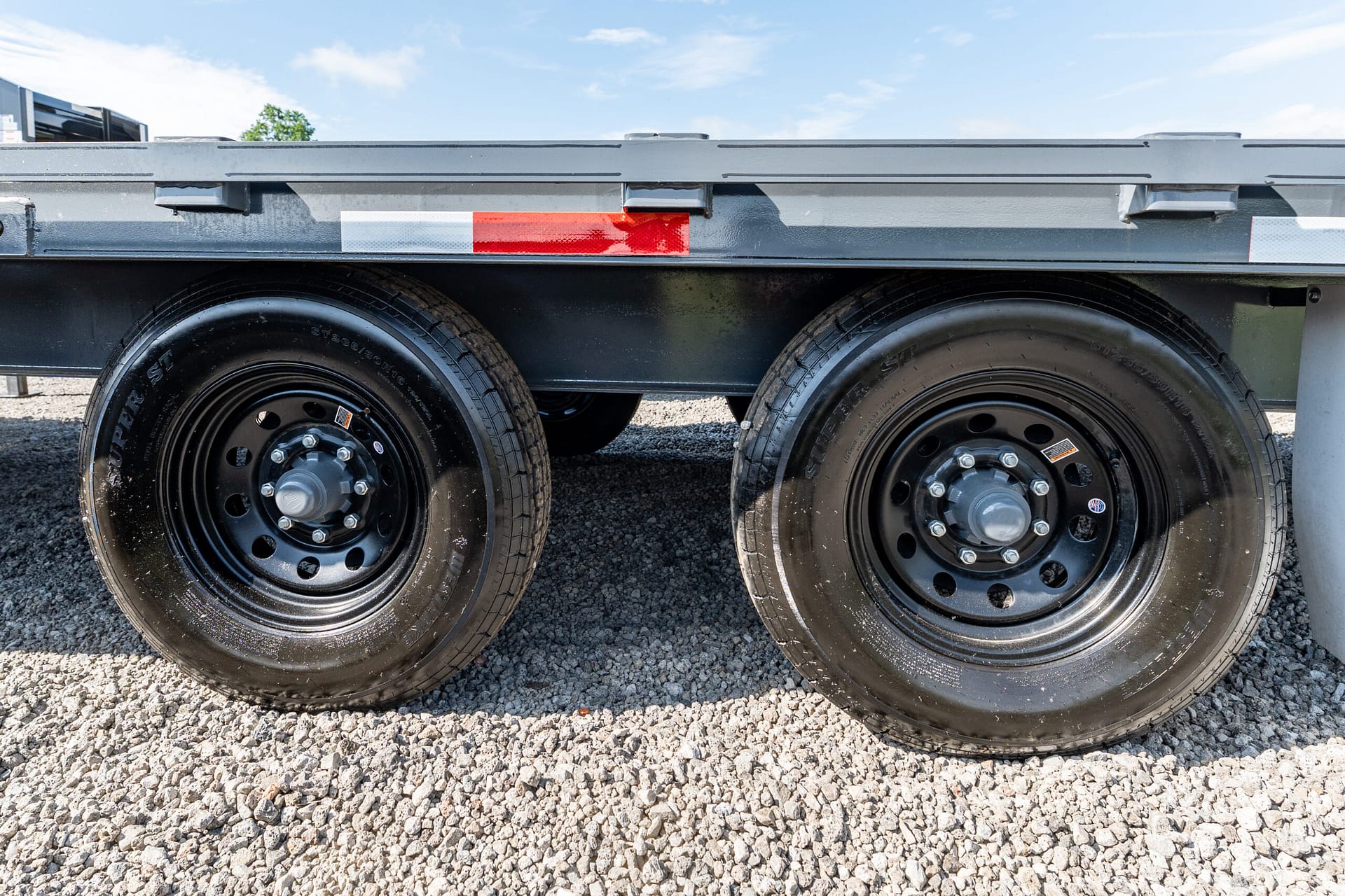 Side view of a trailer axle with two black steel wheels on a gravel surface, blue sky above, and red/white reflective strip nearby.