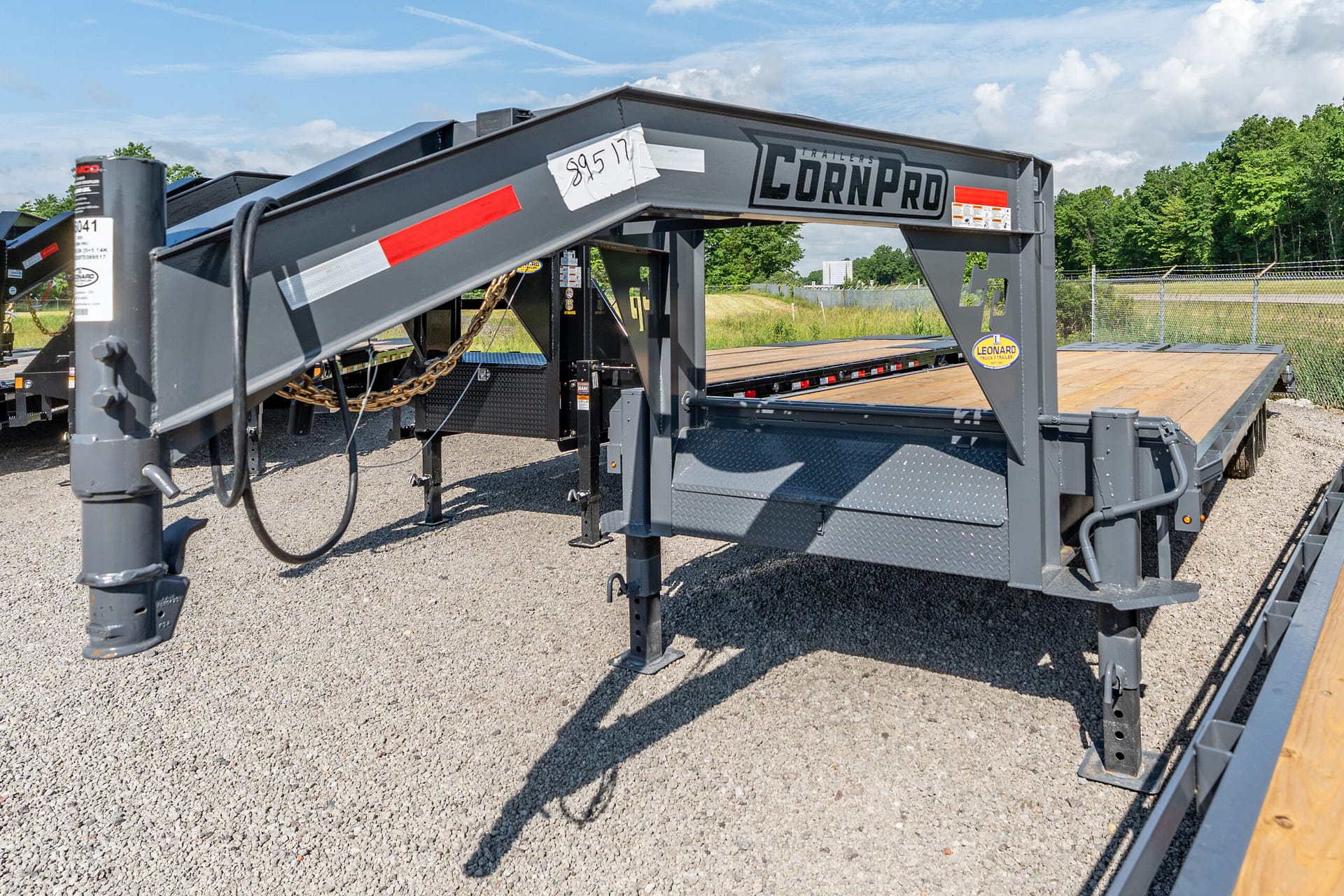 Close-up of a gray CornPro gooseneck trailer with a wooden deck on gravel, safety chains visible, outdoors in a lot.