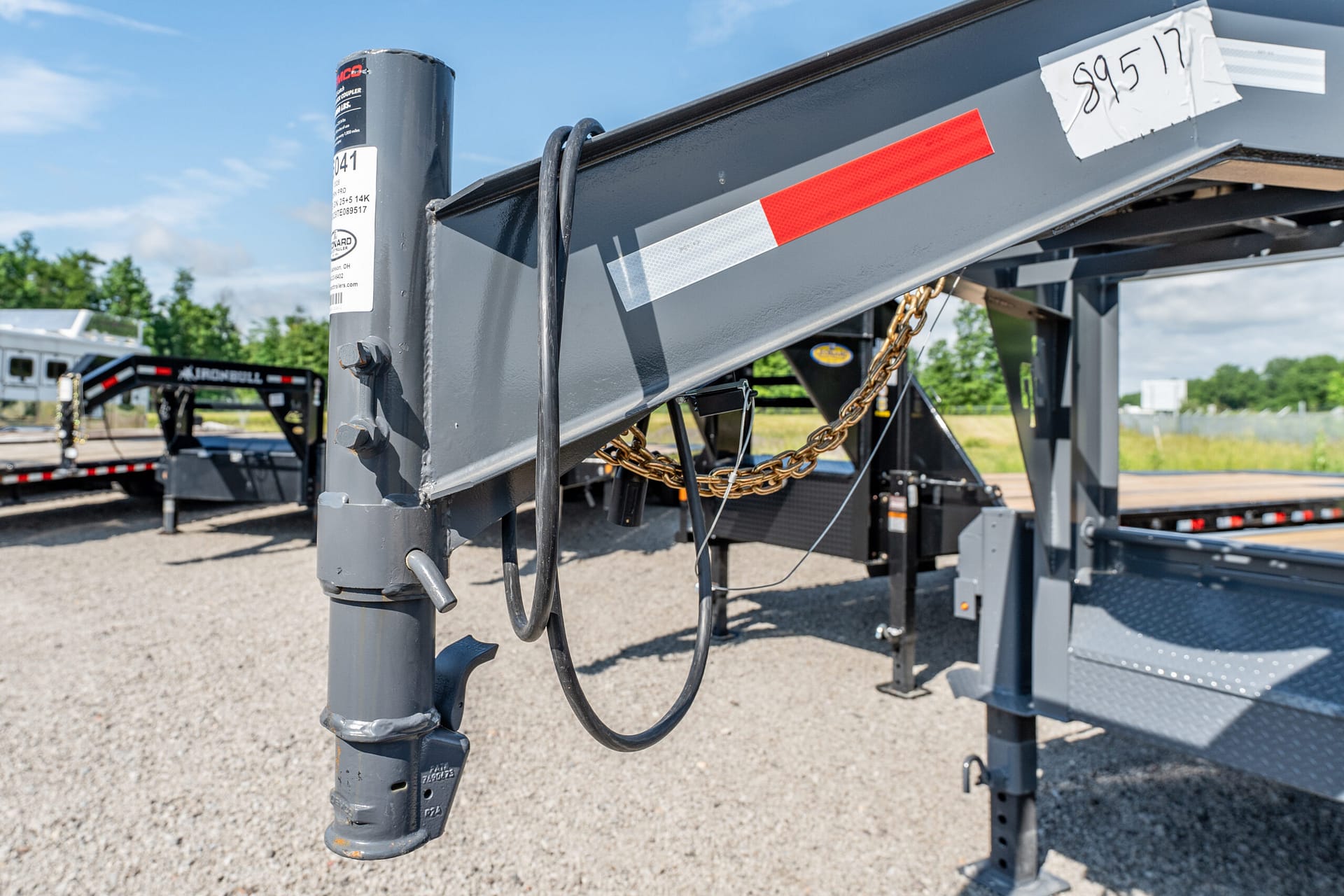Close-up of a gray gooseneck trailer tongue with safety chains and a red reflector stripe in a gravel lot