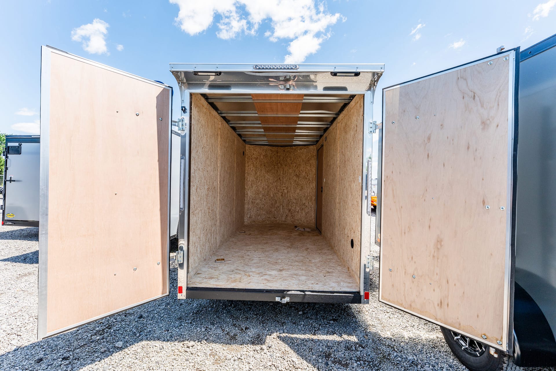 Open empty cargo trailer with plywood interior and double doors swung wide, parked on gravel under a blue sky