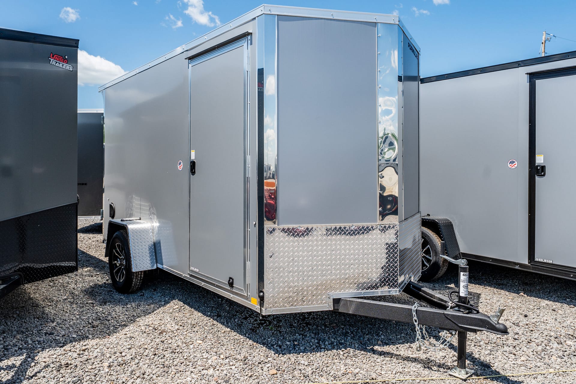 Silver-gray enclosed cargo trailer with diamond-plate front parked on a gravel lot under a blue sky.