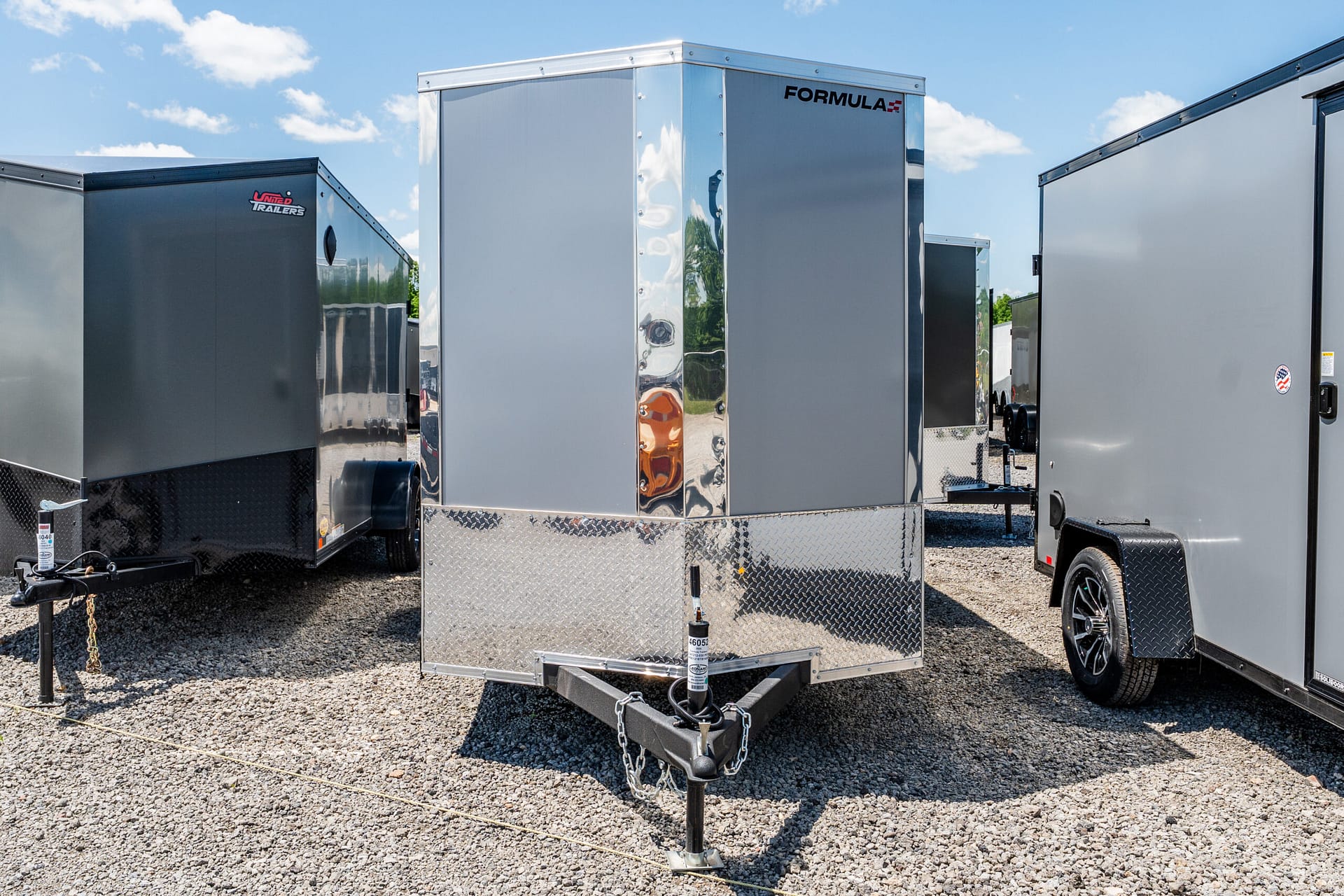 Row of silver and gray enclosed cargo trailers parked on a gravel lot under a bright blue sky with clouds.
