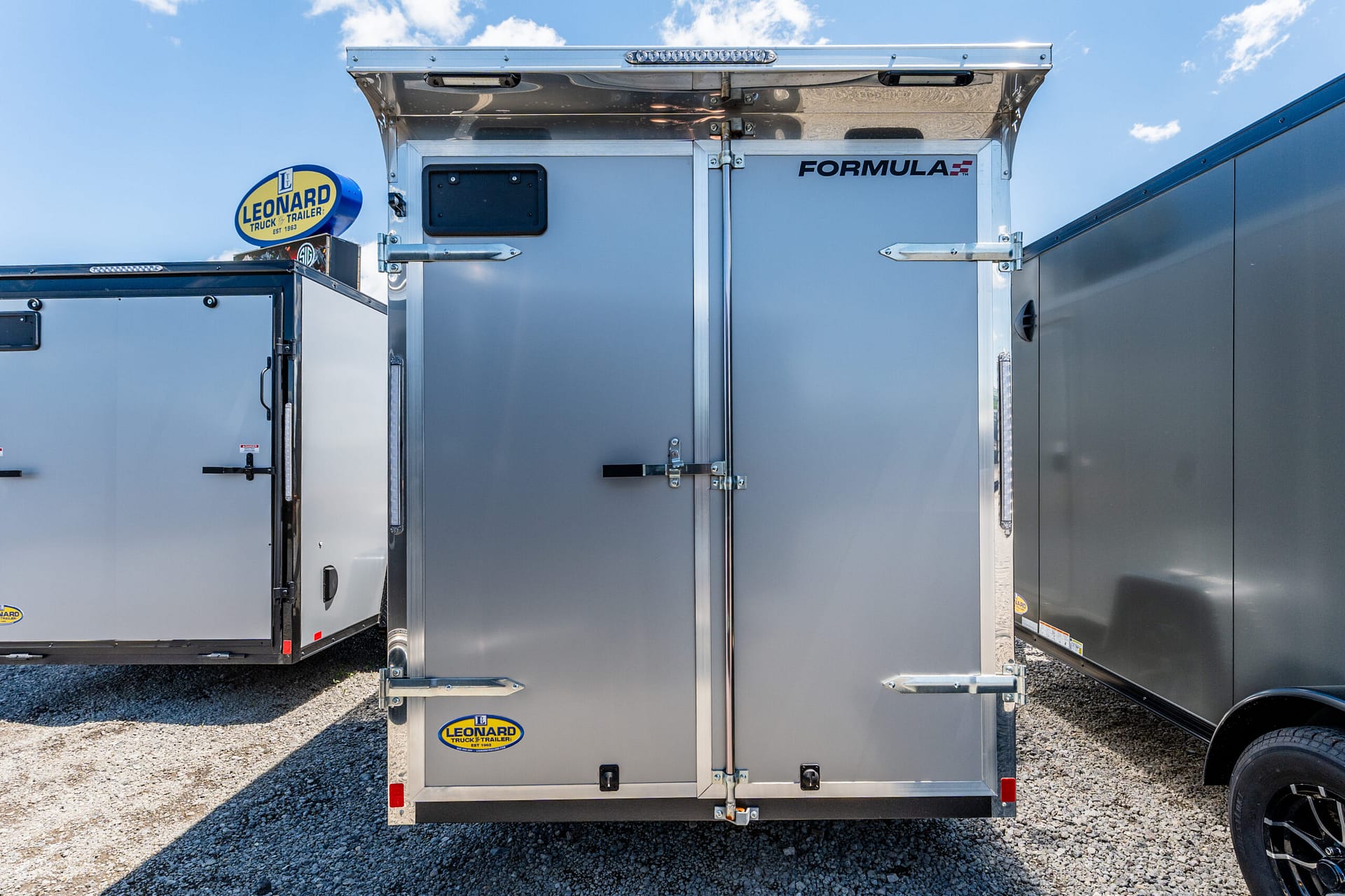 Rear view of a silver cargo trailer with a Leonard Truck & Trailer sticker on the door, against a clear blue sky