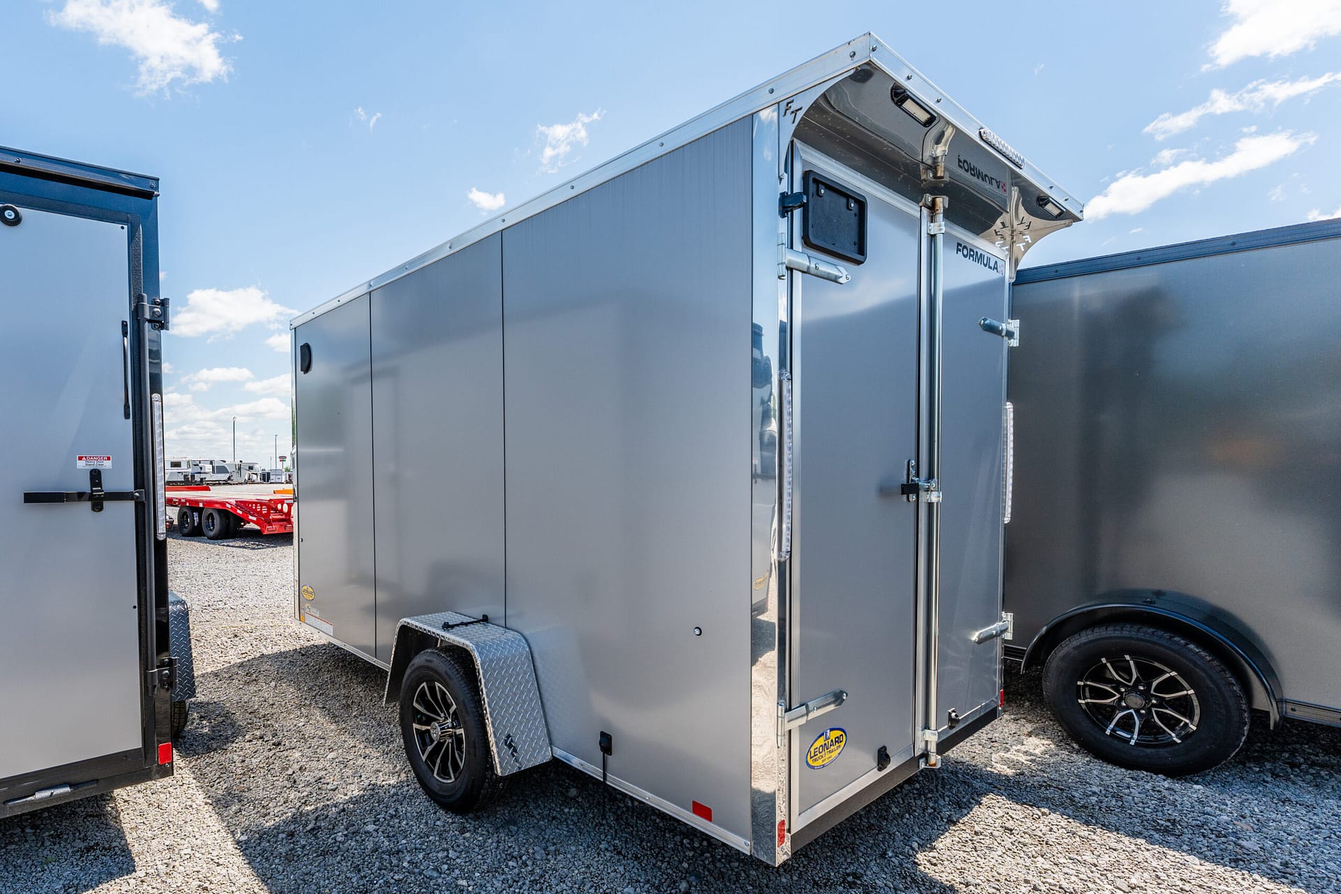 Gray enclosed cargo trailer with a back ramp door, parked on gravel under a blue sky.