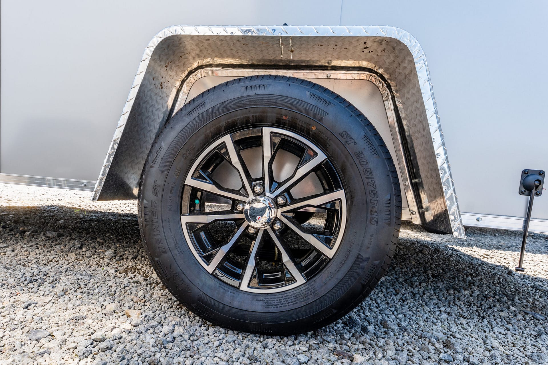 Close-up of a rugged tire and alloy wheel mounted on a trailer, with a diamond-plate fender above and gravel ground beneath.