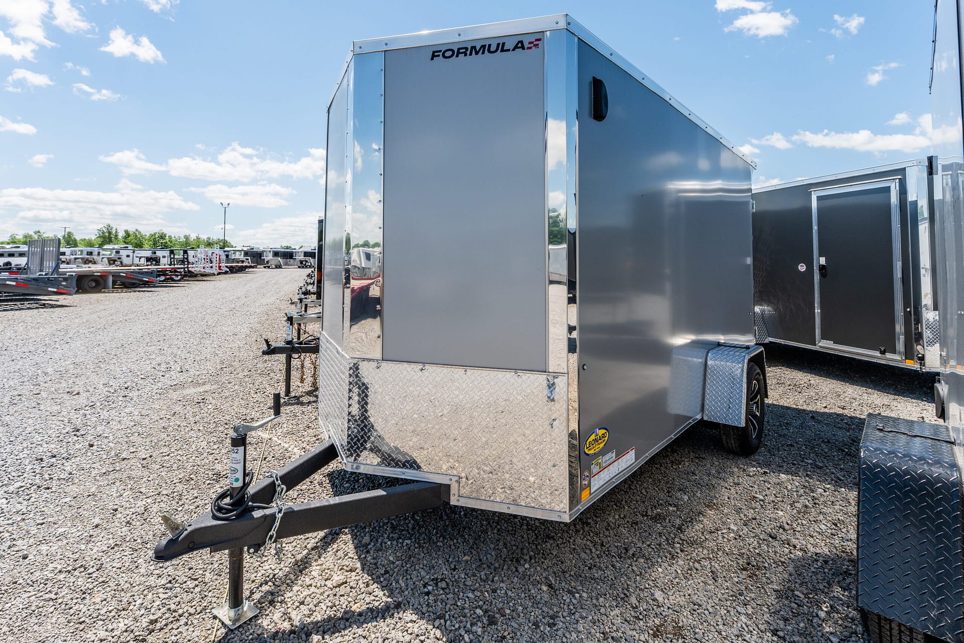 Large silver enclosed trailer at a trailer lot, with rows of trailers in the background and a bright blue sky above.