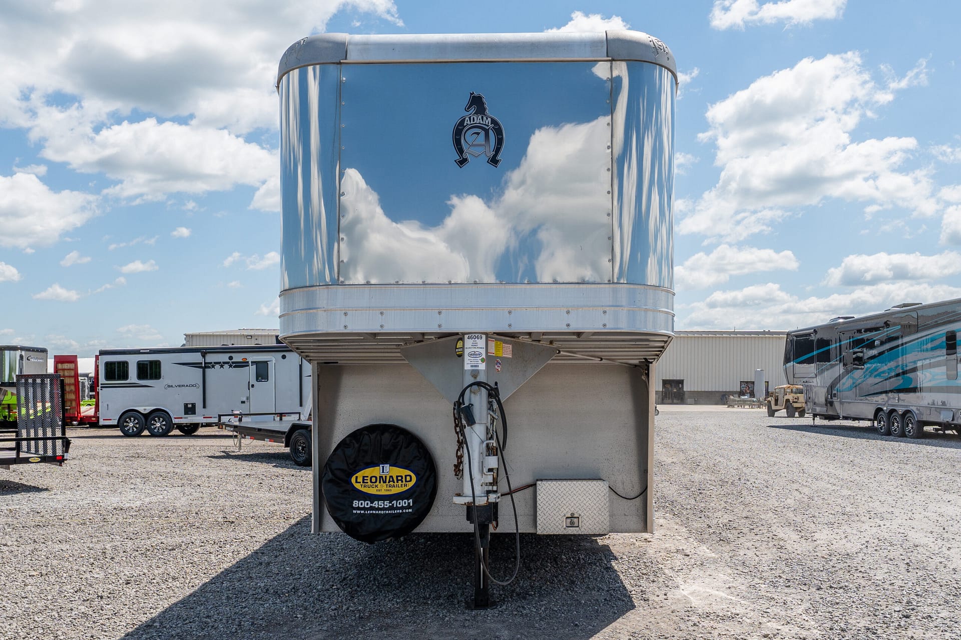 Shiny silver gooseneck trailer front with Leonard Truck & Trailer spare tire cover, parked among other trailers under a blue sky.