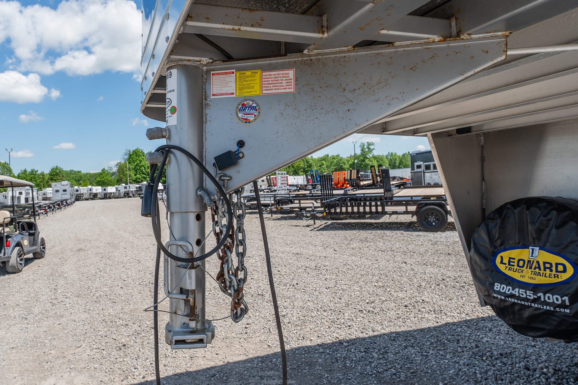 Close-up view of a trailer hitch and weathered steel support beam with rust, chains, and cables hanging down, at a trailer yard under a blue sky with clouds and a Leonard trailer cover visible at right.