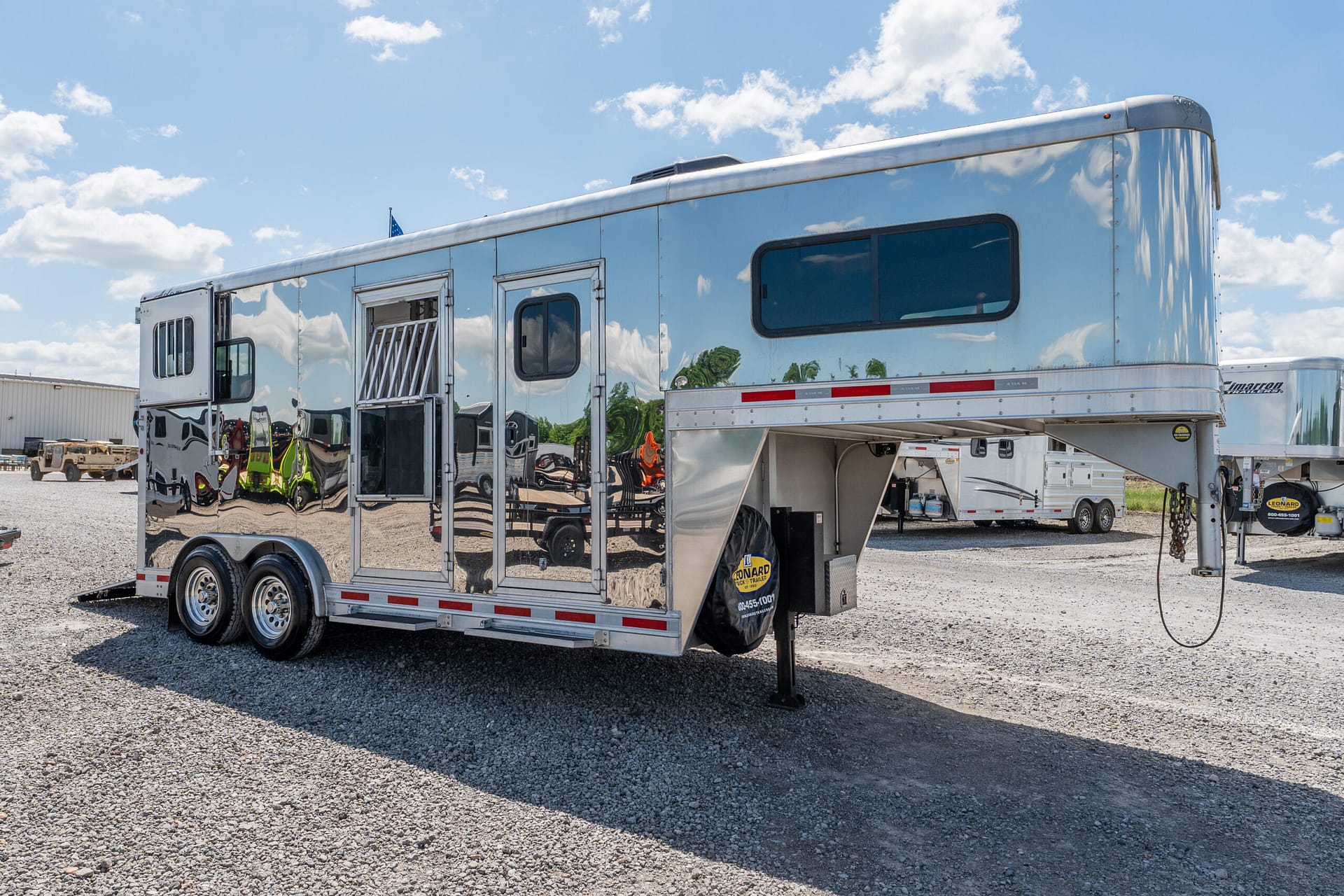 Shiny silver horse trailer with rounded upper front, windows, and a side door parked on a gravel lot with other trailers, under a blue sky.