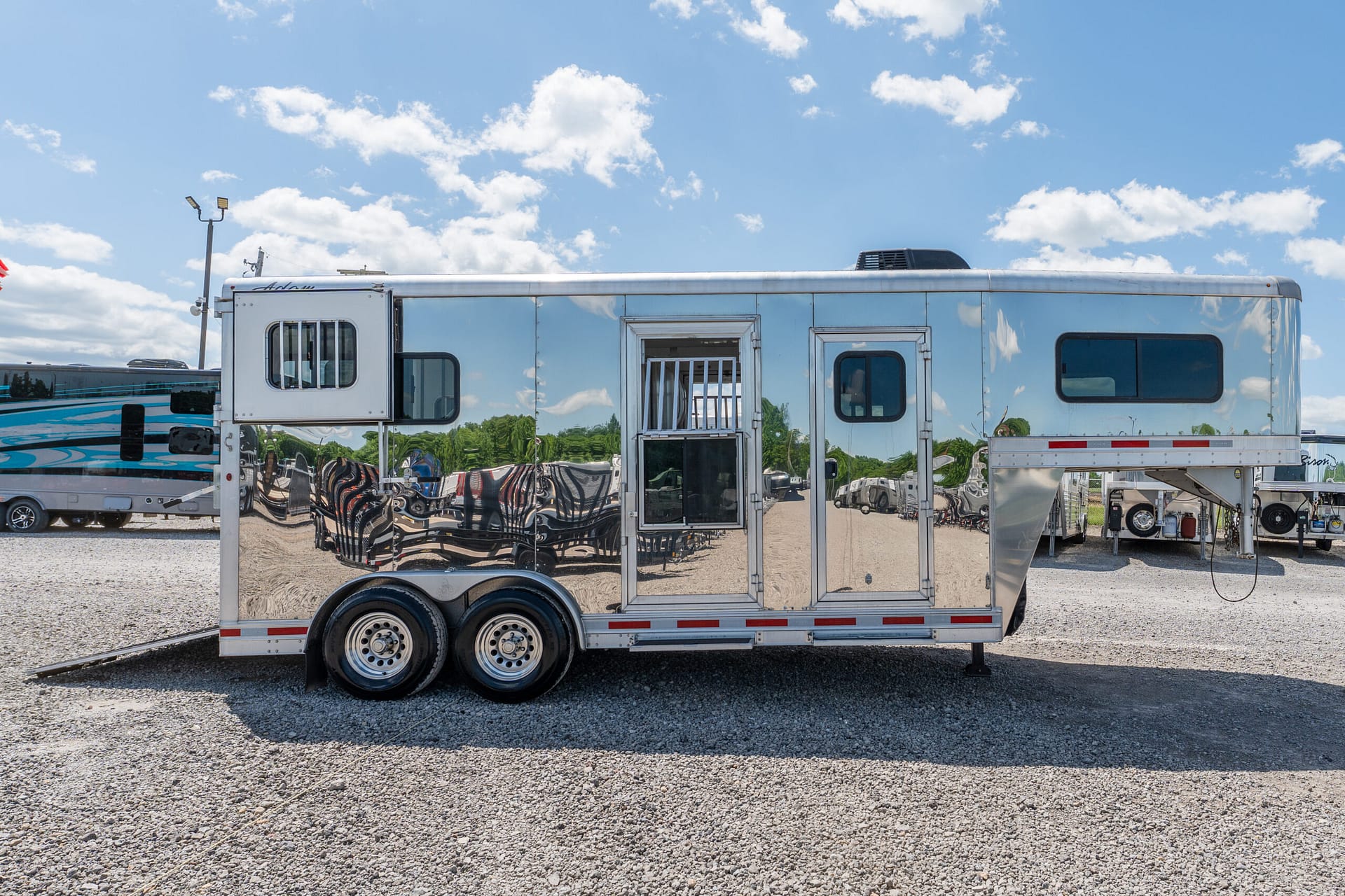 Polished aluminum travel trailer reflecting the surrounding gravel lot, vehicles, and blue sky with clouds.