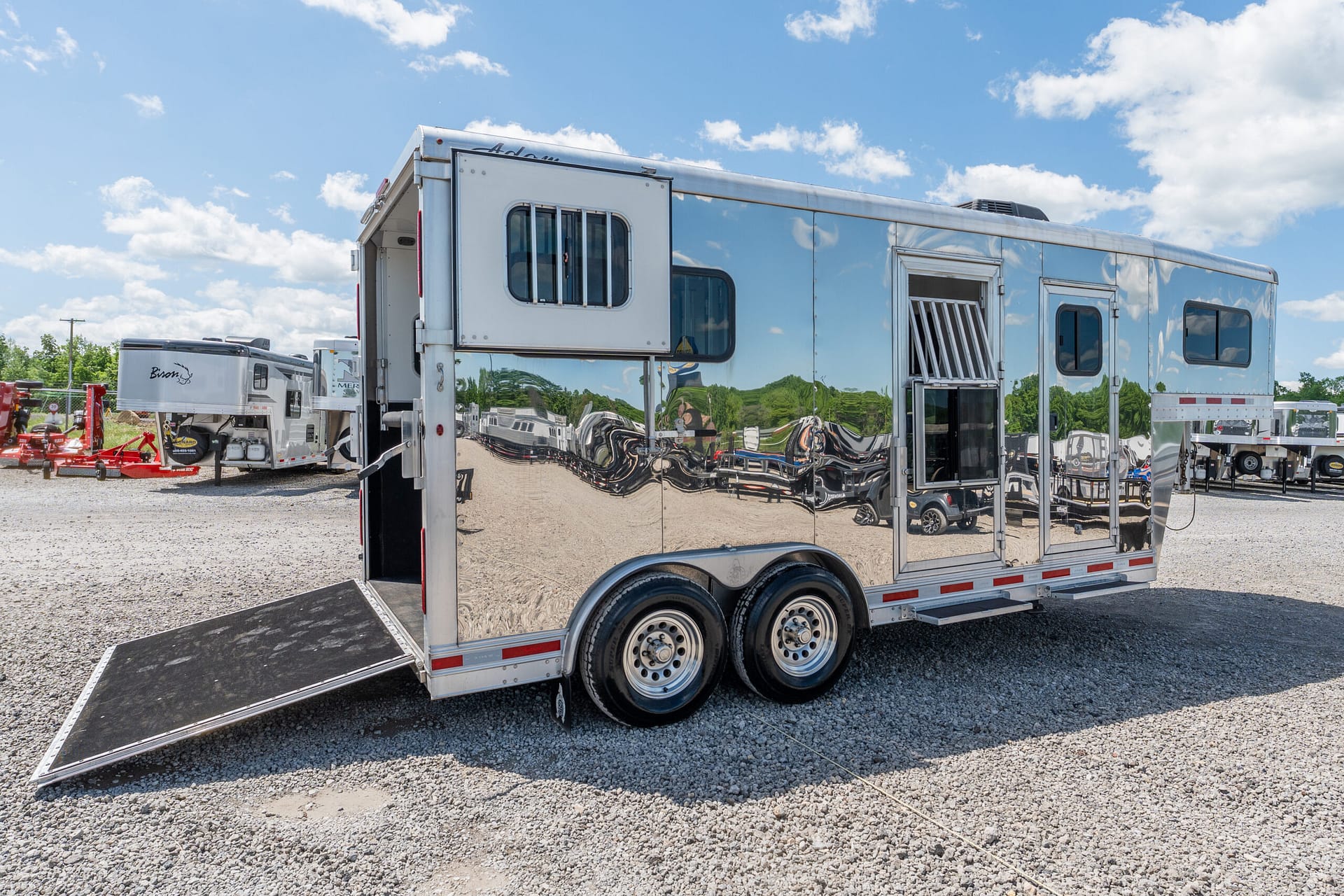 Shiny silver horse trailer with a lowered ramp on a gravel lot, reflections of other trailers and blue sky above.