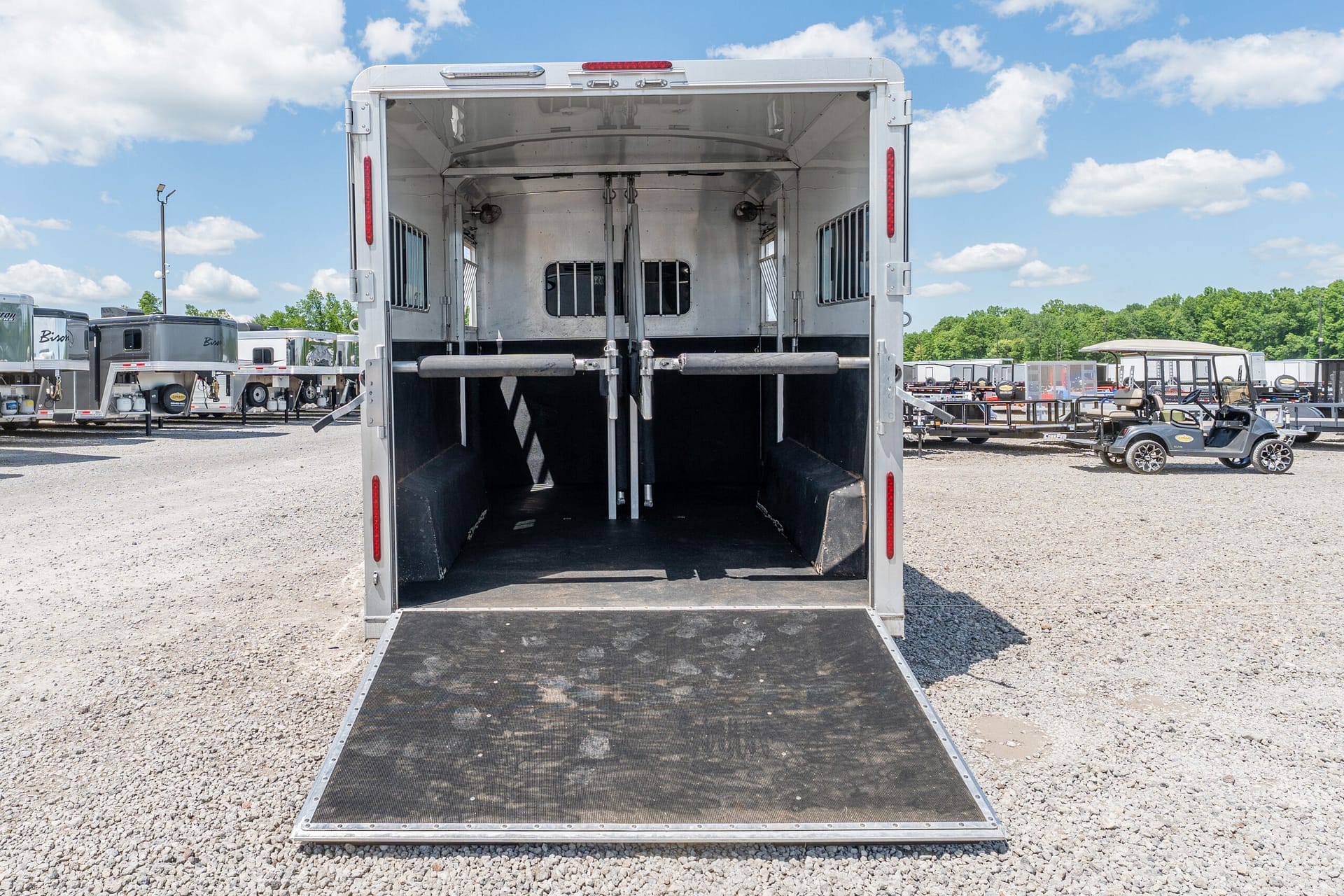 Rear view of an empty horse trailer with stalls and partition, ramp down, in a gravel lot with other trailers in the background under a blue sky.