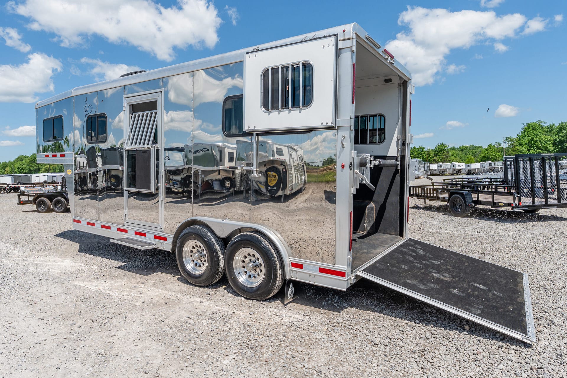Shiny aluminum horse trailer with the rear ramp lowered on a gravel lot under a blue sky with clouds