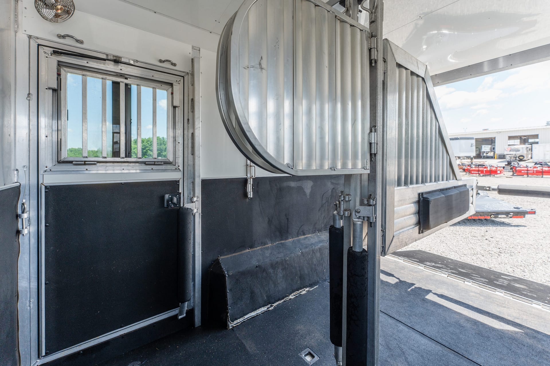 Interior of a silver trailer with metal doors and a curved, corrugated ramp near the entrance; outdoors shows trucks and a blue sky.