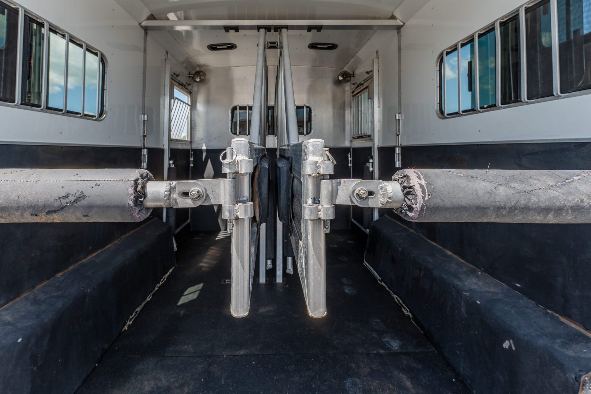 Interior of a livestock trailer with central metal dividers, rubber-matted floor, and side windows, viewed from the rear.