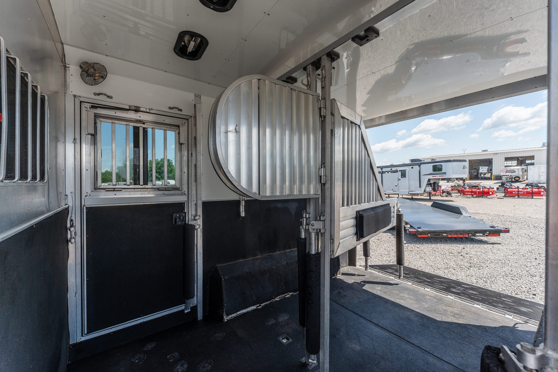 Interior of a shiny aluminum trailer with metal stall dividers and a window, outdoor trailer yard visible through the open door far end.