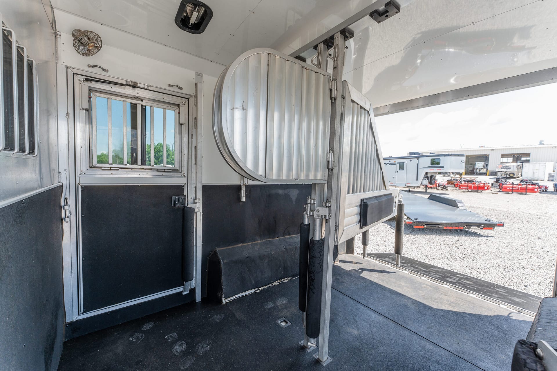 Interior of a shiny aluminum horse trailer with a curved metal divider and barred window on the left; other stalls and outside equipment visible through the open back.