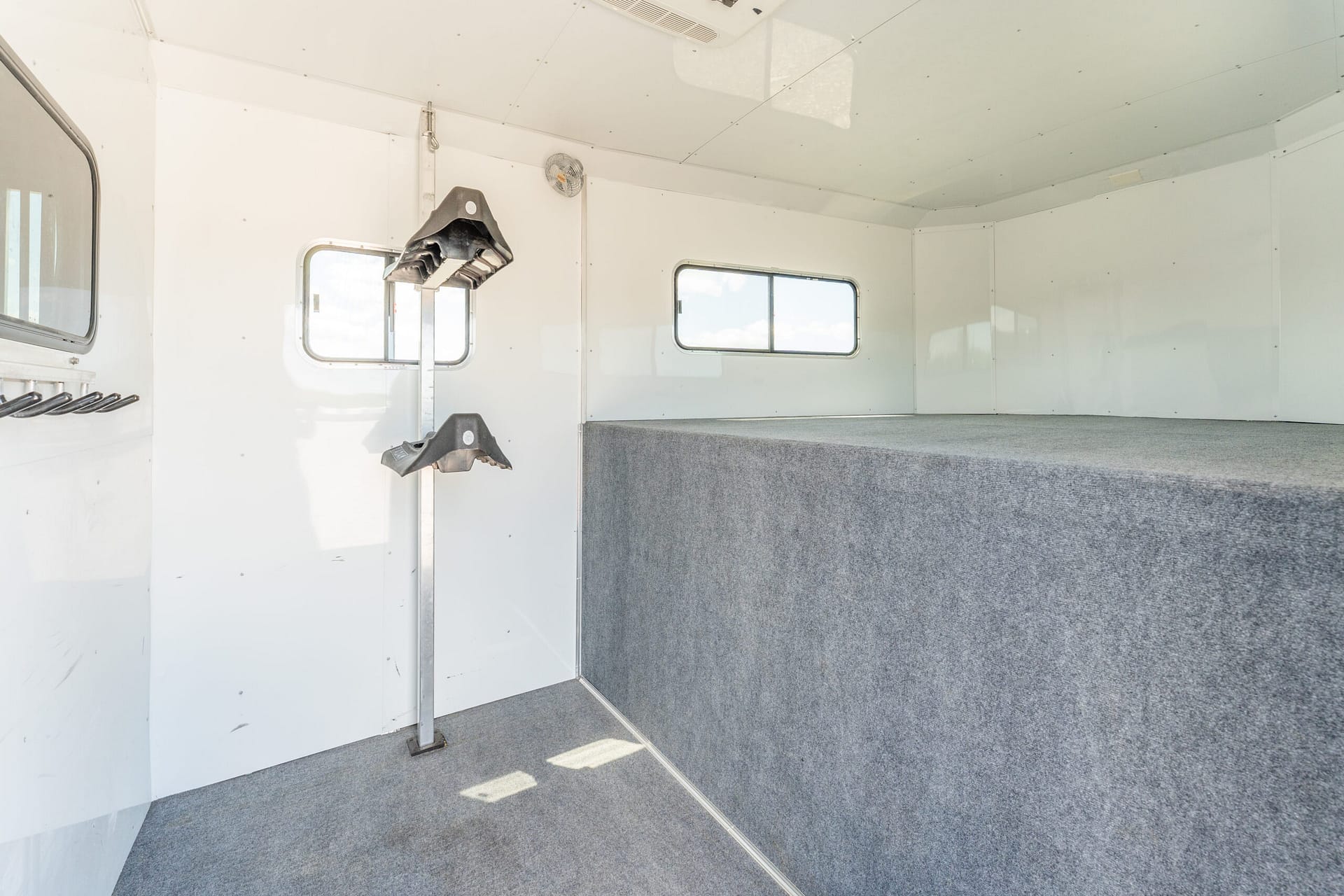 Interior of a white trailer with gray carpet, featuring wall-mounted black storage hooks and two small windows.