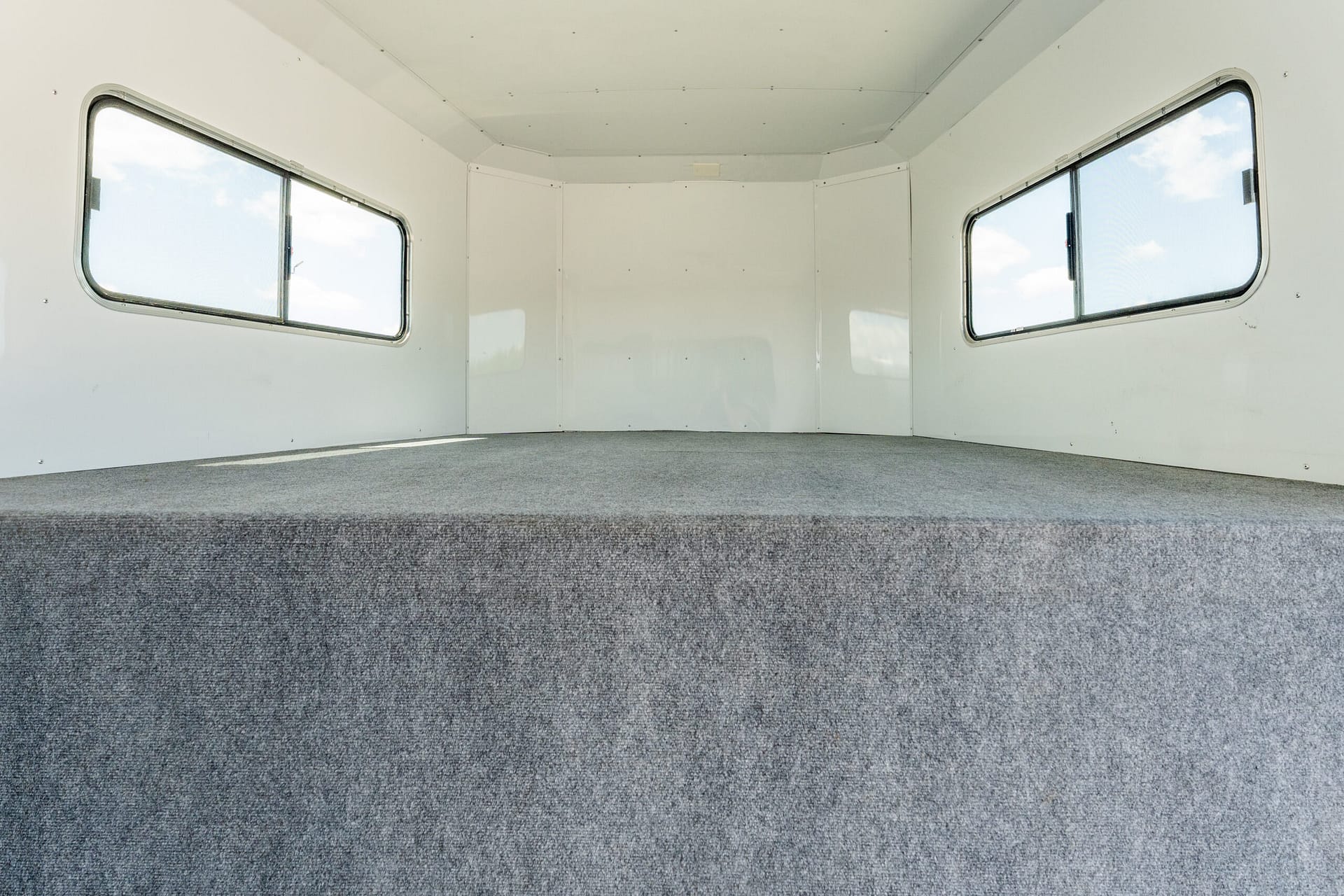 Empty white trailer interior with two large side windows and gray carpeted floor, viewed from the back looking forward.