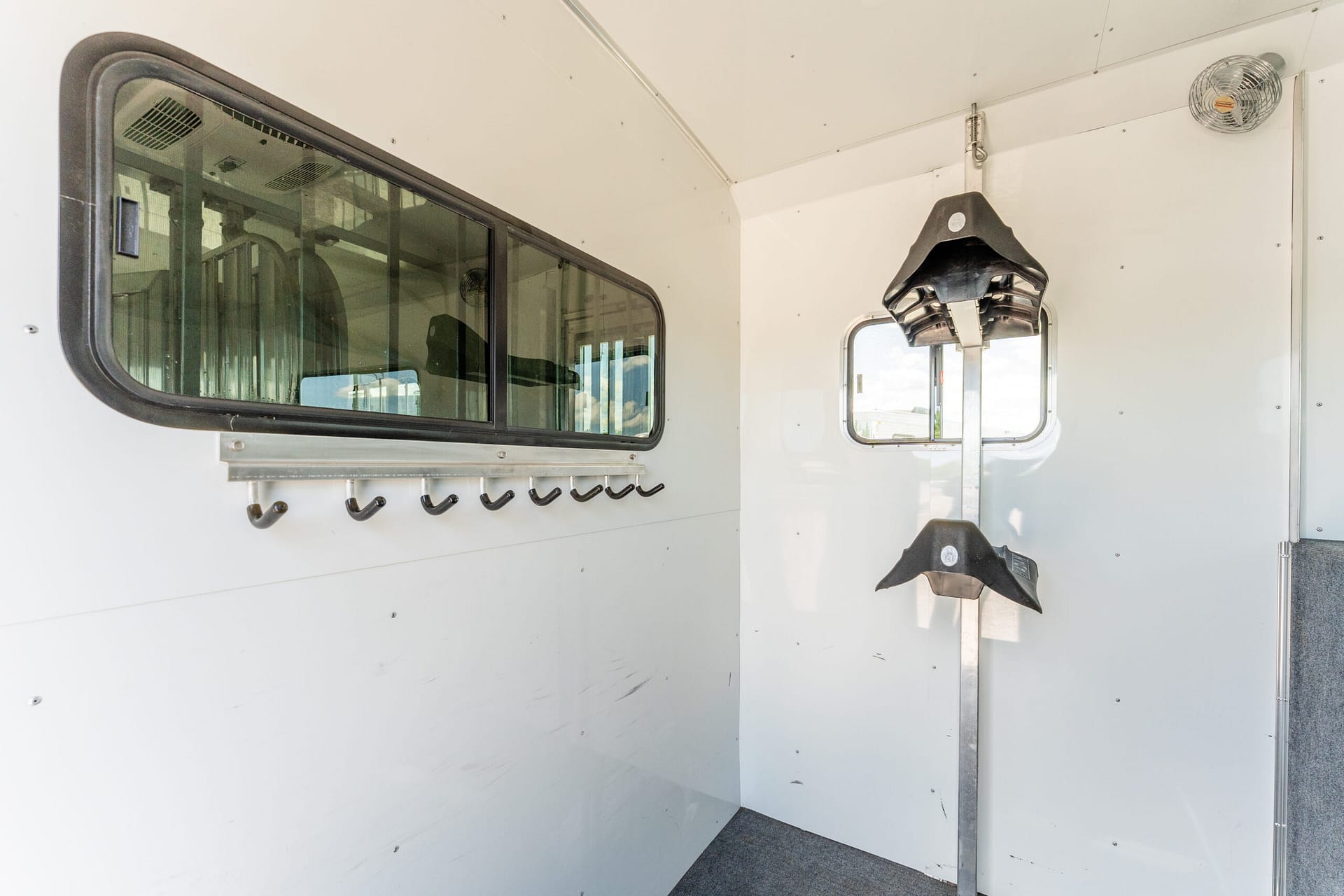 Interior of a white utility trailer showing a large rounded window on the left, row of black hooks beneath, and a vertical rack with dark saddle-like hangers near a small window on the right.